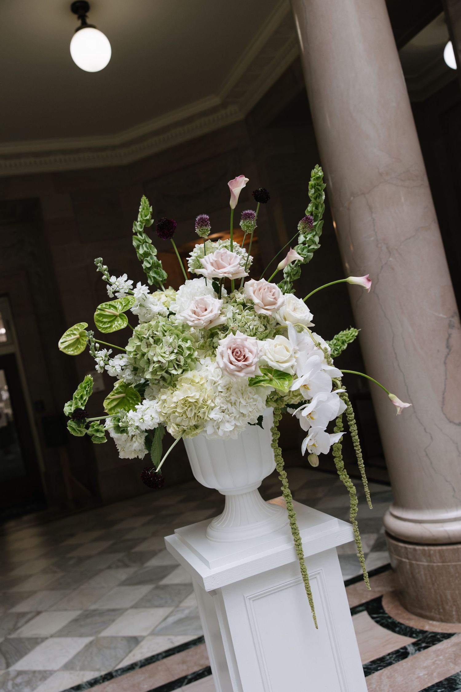 Elegant white urn vase with lush floral arrangement of pink roses, hydrangeas, and greenery on a pedestal in a grand hall.