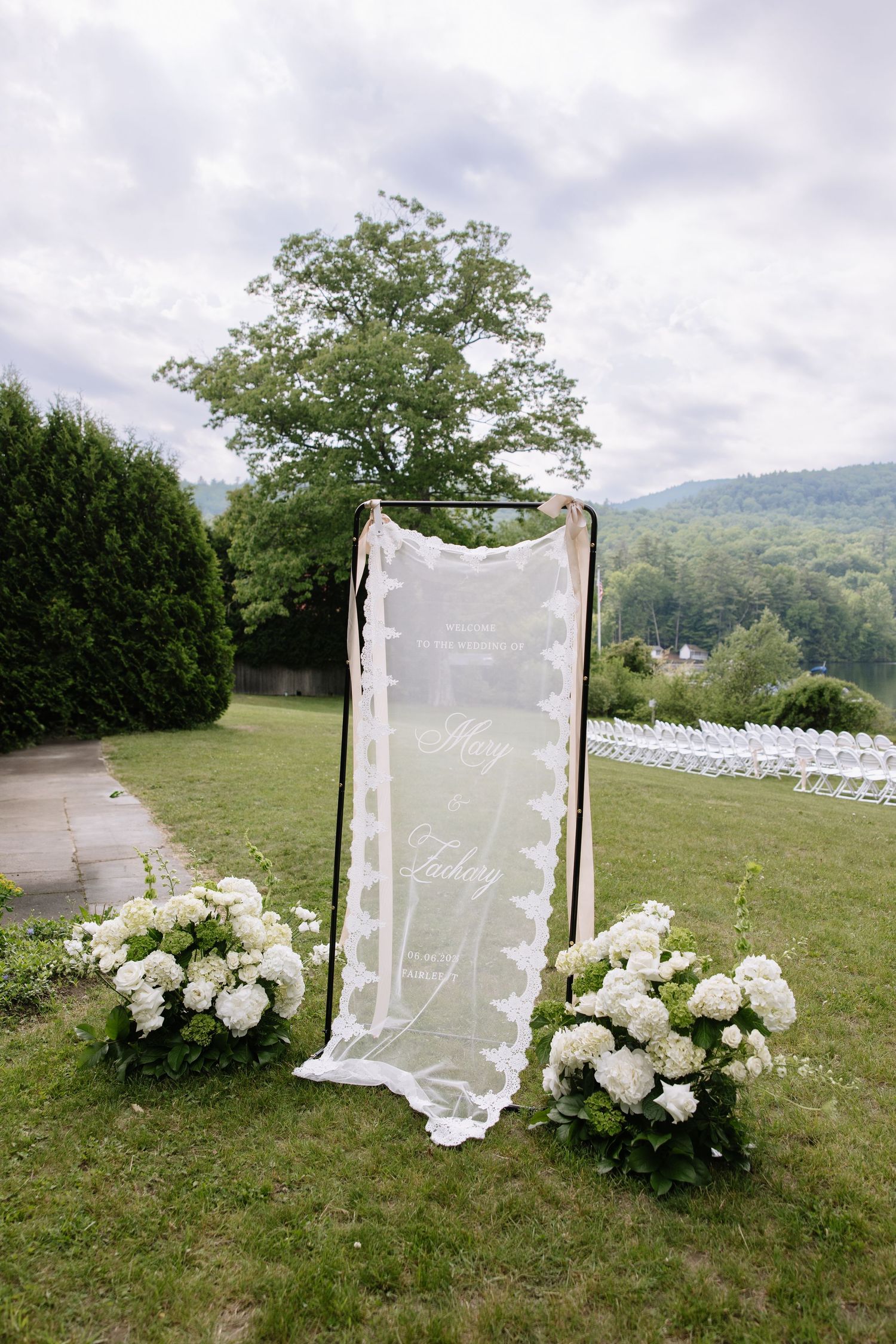 Outdoor wedding ceremony setup with white floral arrangements and an elegant sheer veil sign on a black frame arch.