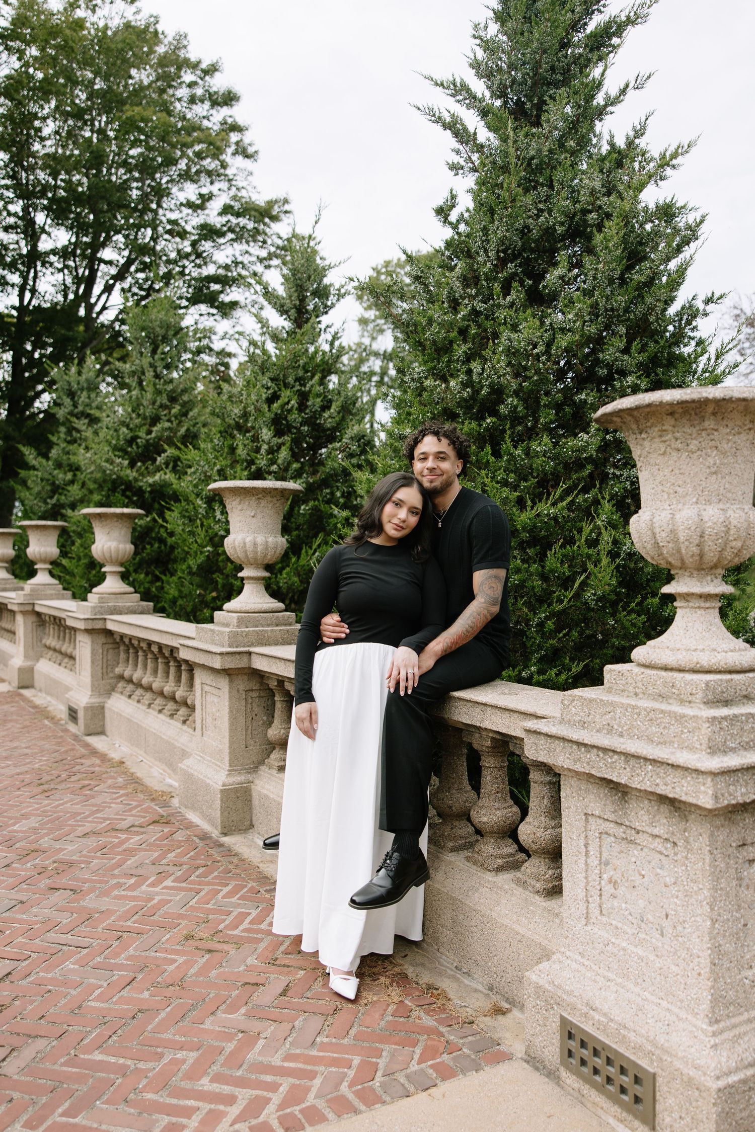 Couple posing on ornate stone balustrade with evergreen trees in background.
