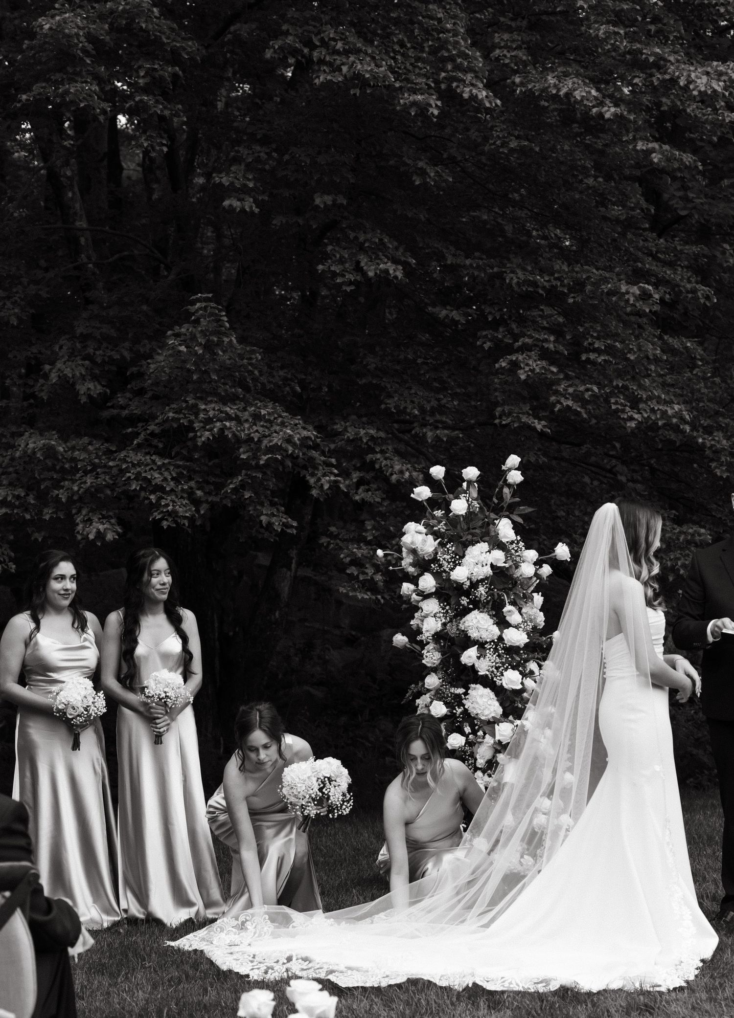 Bridesmaids in silk dresses attend to the bride's long veil during an outdoor ceremony.