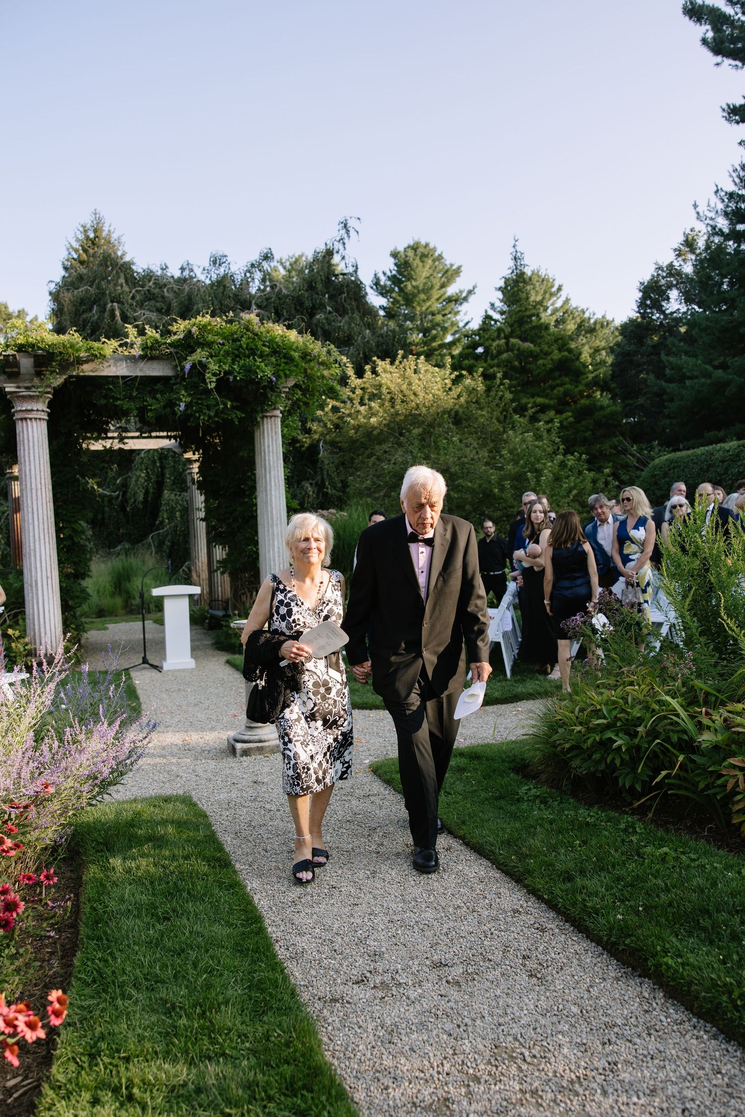 Wedding guests stroll along a scenic gravel path lined with gardens and decorative columns at an outdoor venue.
