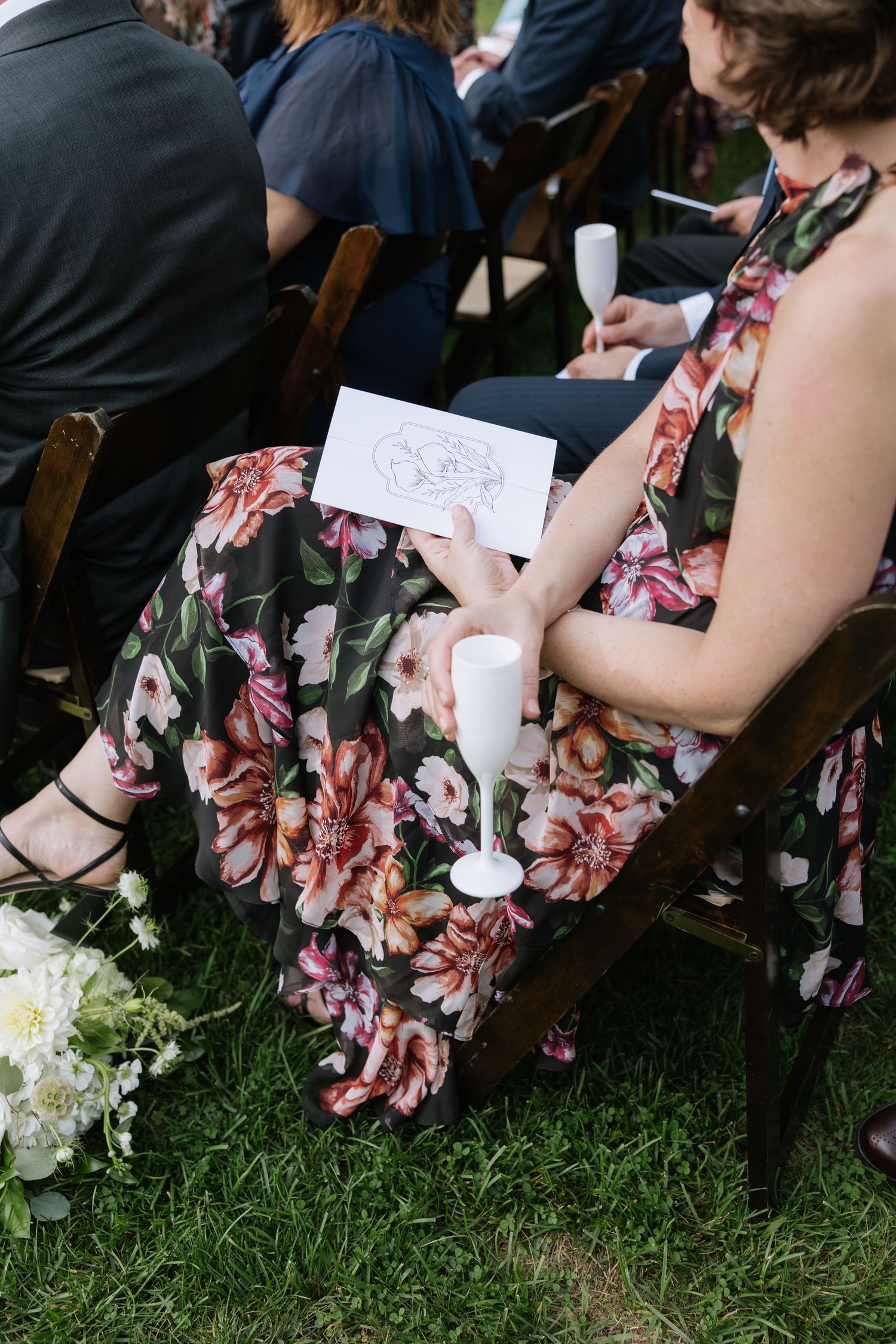 Wedding guests in floral dresses seated on chairs holding ceremony programs.