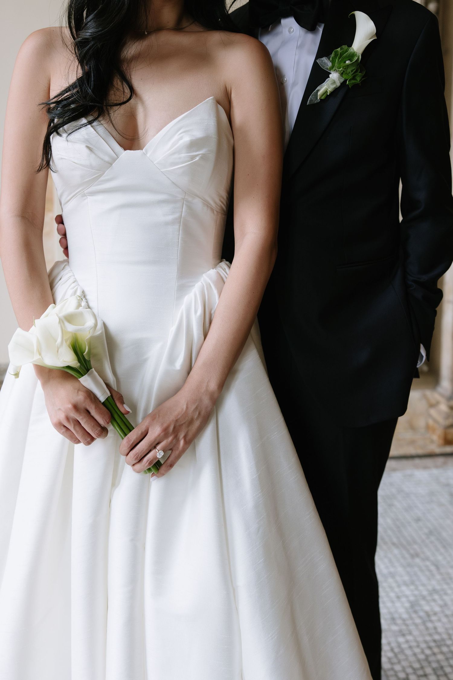 Bride in strapless white gown holding calla lily bouquet stands beside groom in black tuxedo at wedding ceremony.