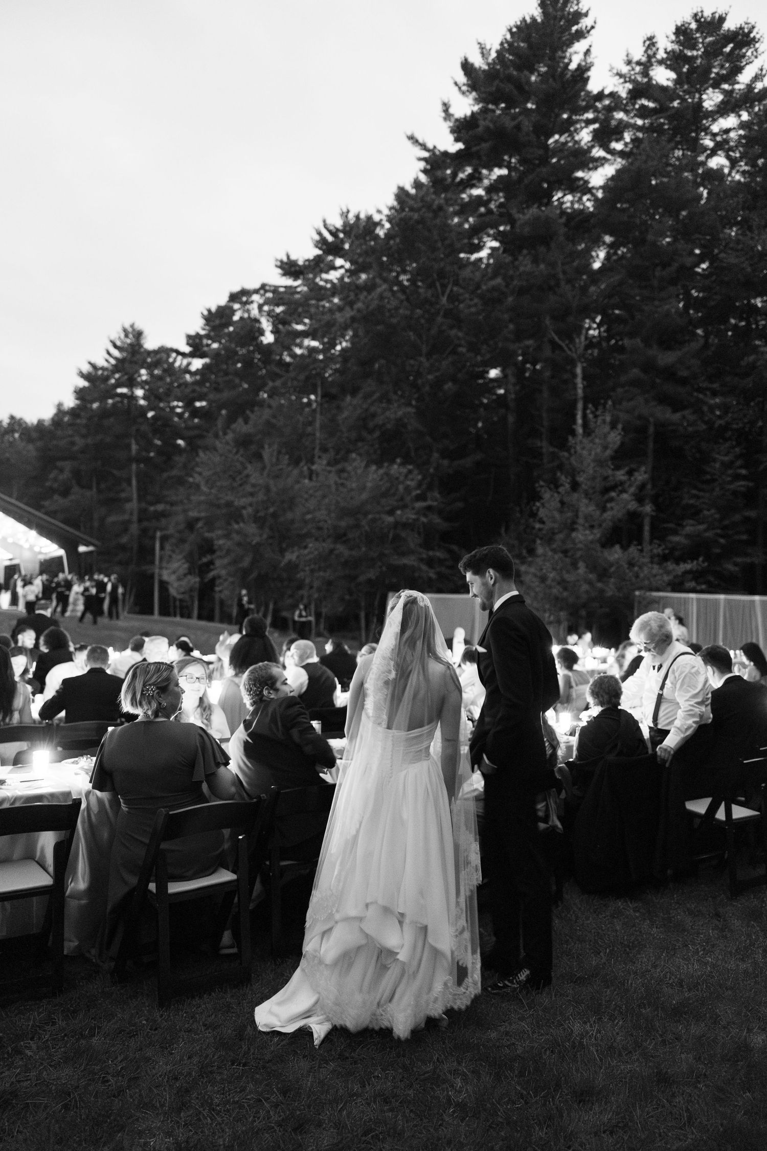 Black and white photo of bride and groom walking among outdoor wedding reception tables with tall pine trees in background.