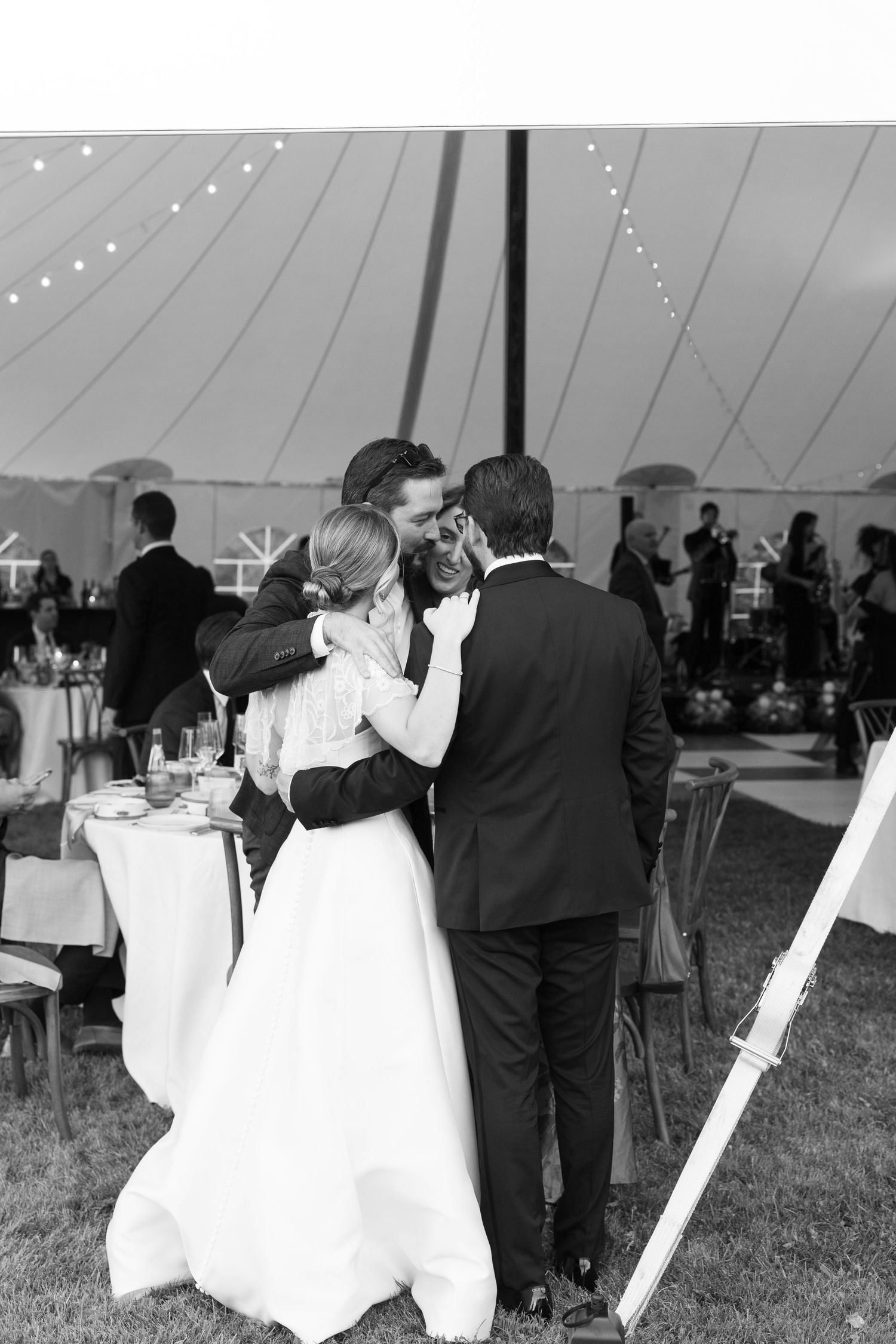 Black and white photo of bride and groom sharing an emotional group hug under a tent at their wedding reception.