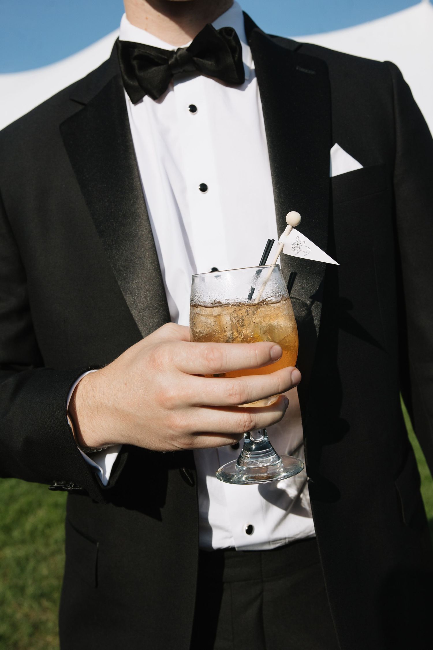Man in black tuxedo with bow tie holding an iced cocktail garnished with a decorative umbrella at a formal event.