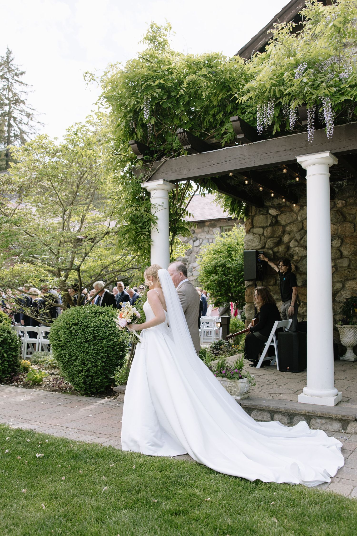Bride in white gown with long veil walks toward outdoor wedding ceremony at stone venue with wisteria-draped pergola.