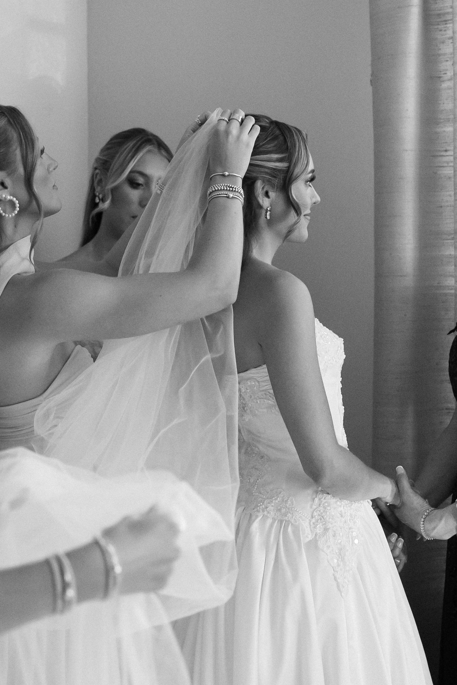 Bridesmaids helping bride put on veil before wedding ceremony in black and white photo.
