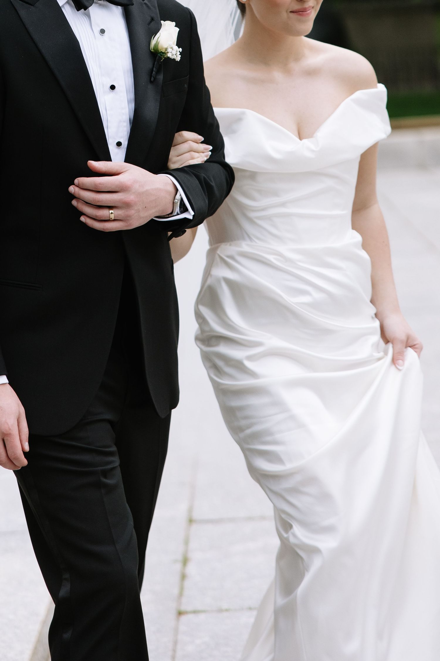 Bride in off-shoulder white gown walks arm-in-arm with groom in black tuxedo on stone pathway.
