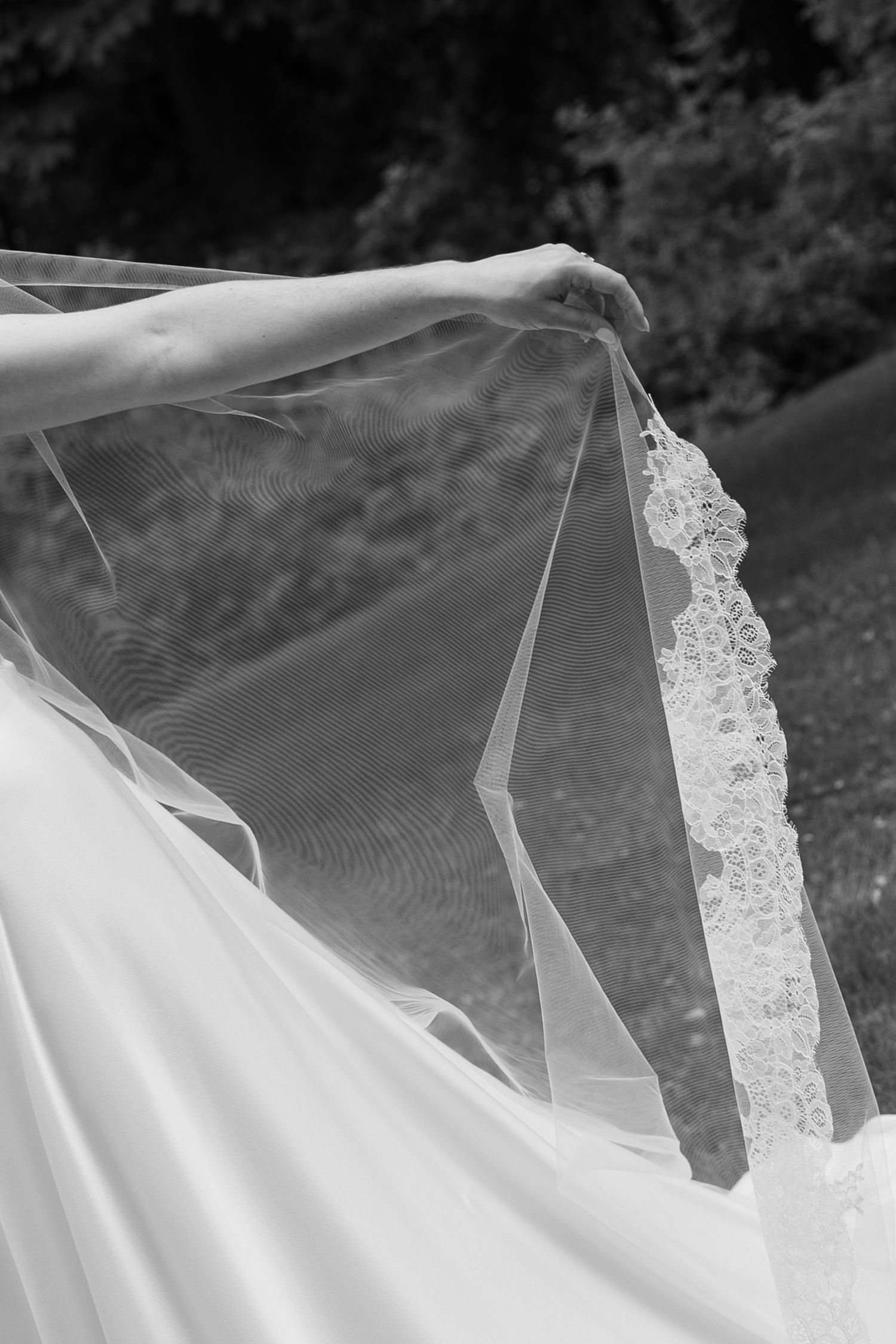 Black and white photo of a bride holding her lace-trimmed wedding veil outdoors.
