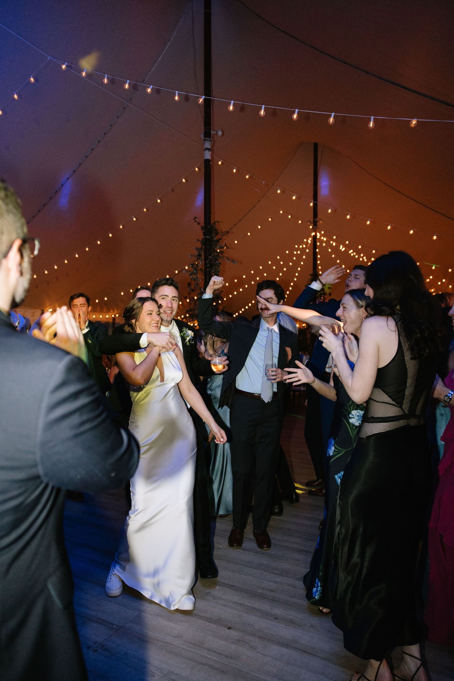 Bride and groom dancing joyfully with guests under string lights in a warm amber wedding tent.
