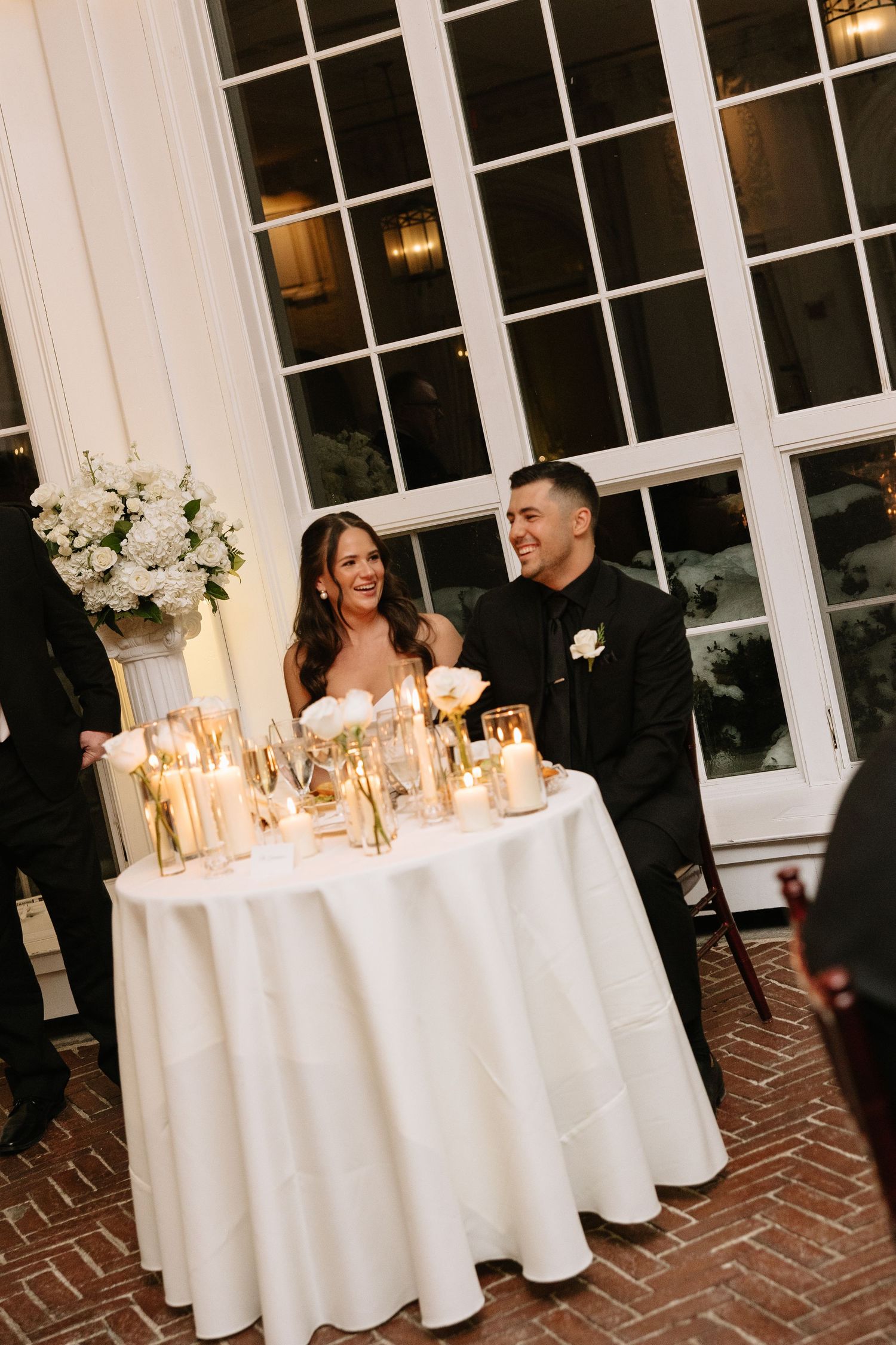 Bride and groom laugh at sweetheart table adorned with gold candles and white florals during elegant wedding reception.