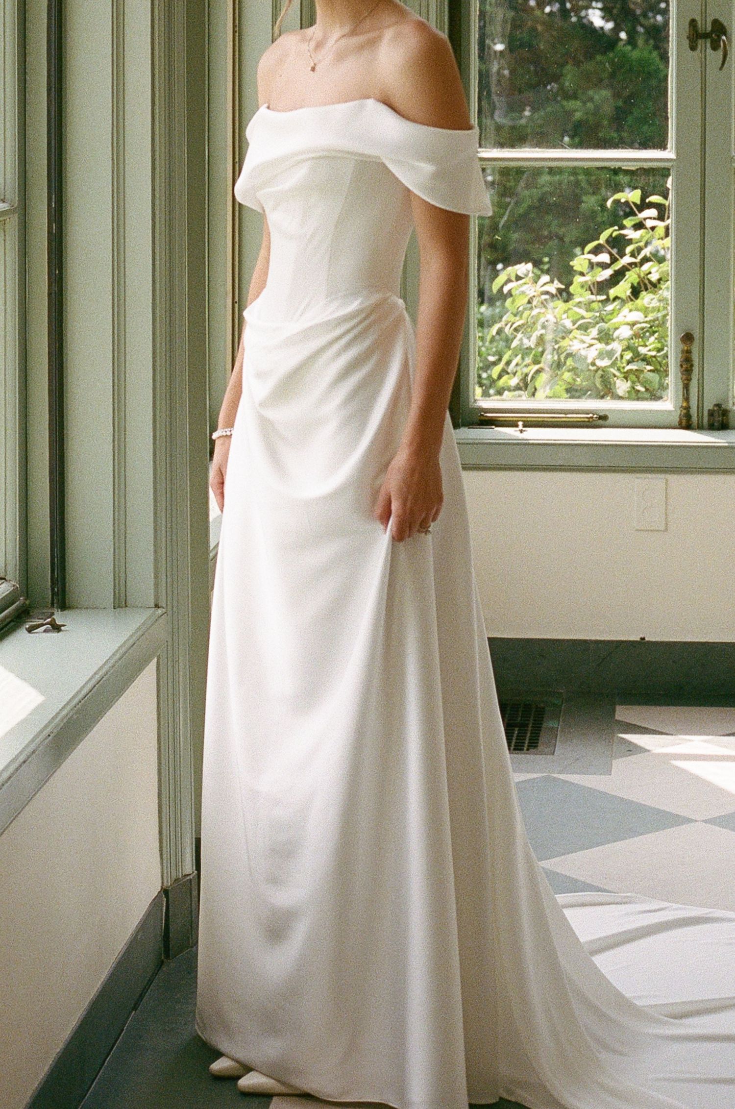 A bride stands beside a sunlit window in an elegant off-the-shoulder satin wedding gown with a long flowing train.