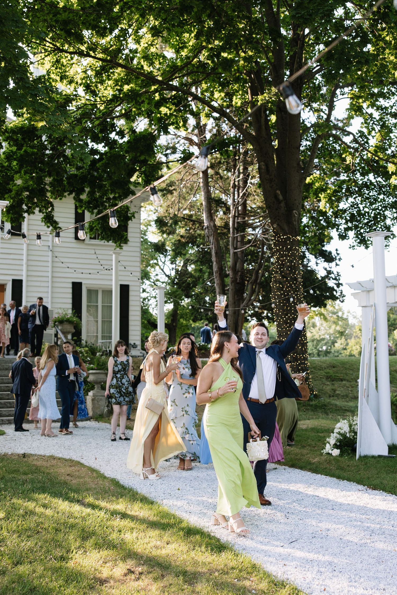 Guests celebrate outdoors at an elegant wedding reception on a gravel path surrounded by lush trees and string lights.