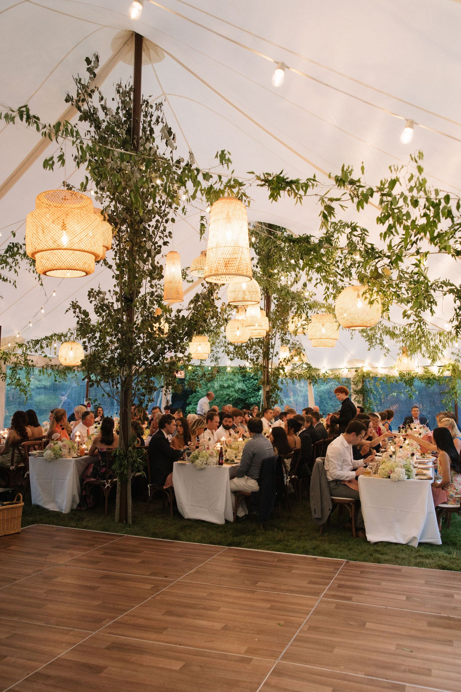 Elegant tented wedding reception with wicker pendant lights, lush greenery, string lights, and guests dining at white-clothed tables.