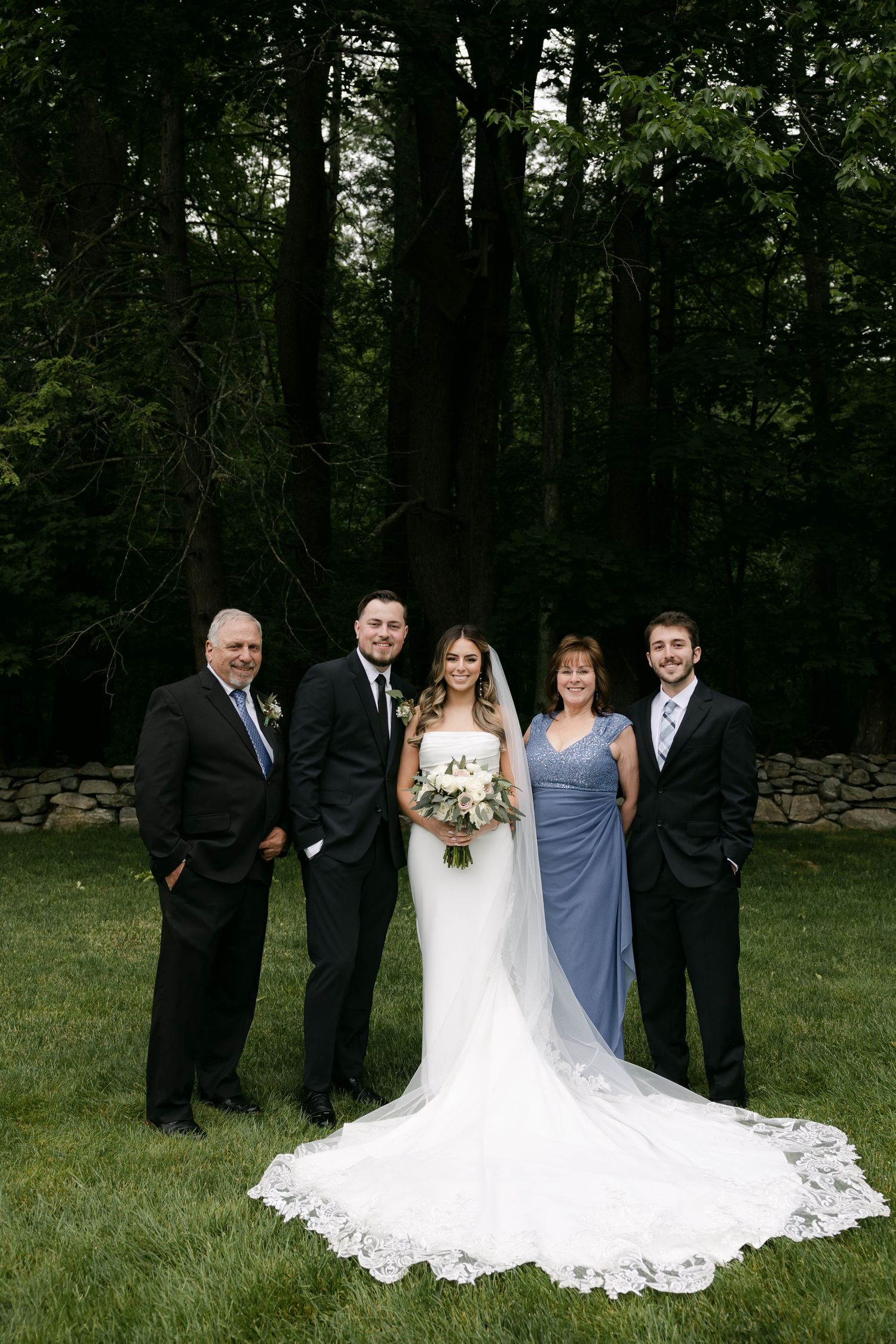 Wedding party gathered for formal photos on lawn with stone wall and forest backdrop.