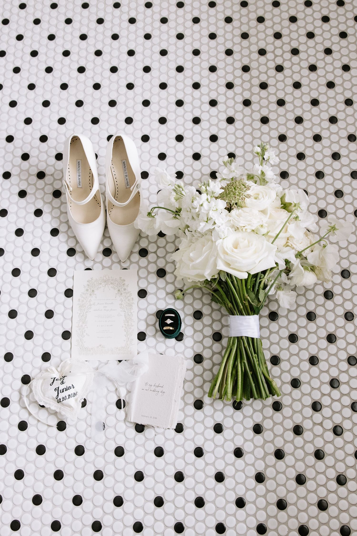 Wedding details flatlay with white shoes, floral bouquet, and stationery on polka dot tile background.