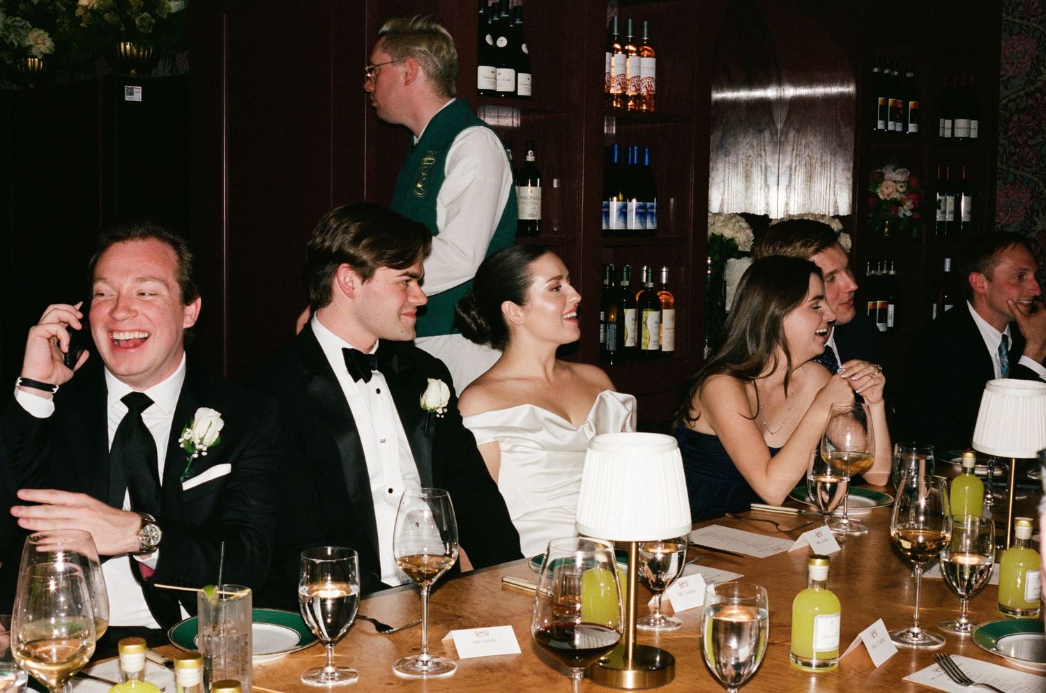 Wedding reception guests laughing and celebrating at a formal dinner table with wine glasses and floral centerpieces.