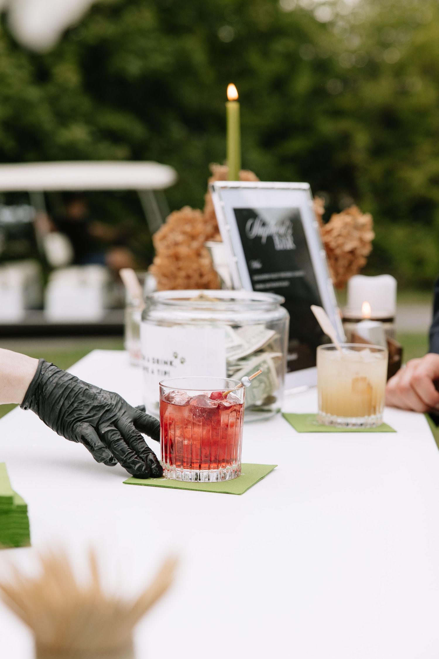 Bartender in black gloves serving a red cocktail on a white outdoor bar table with candles and drinks.