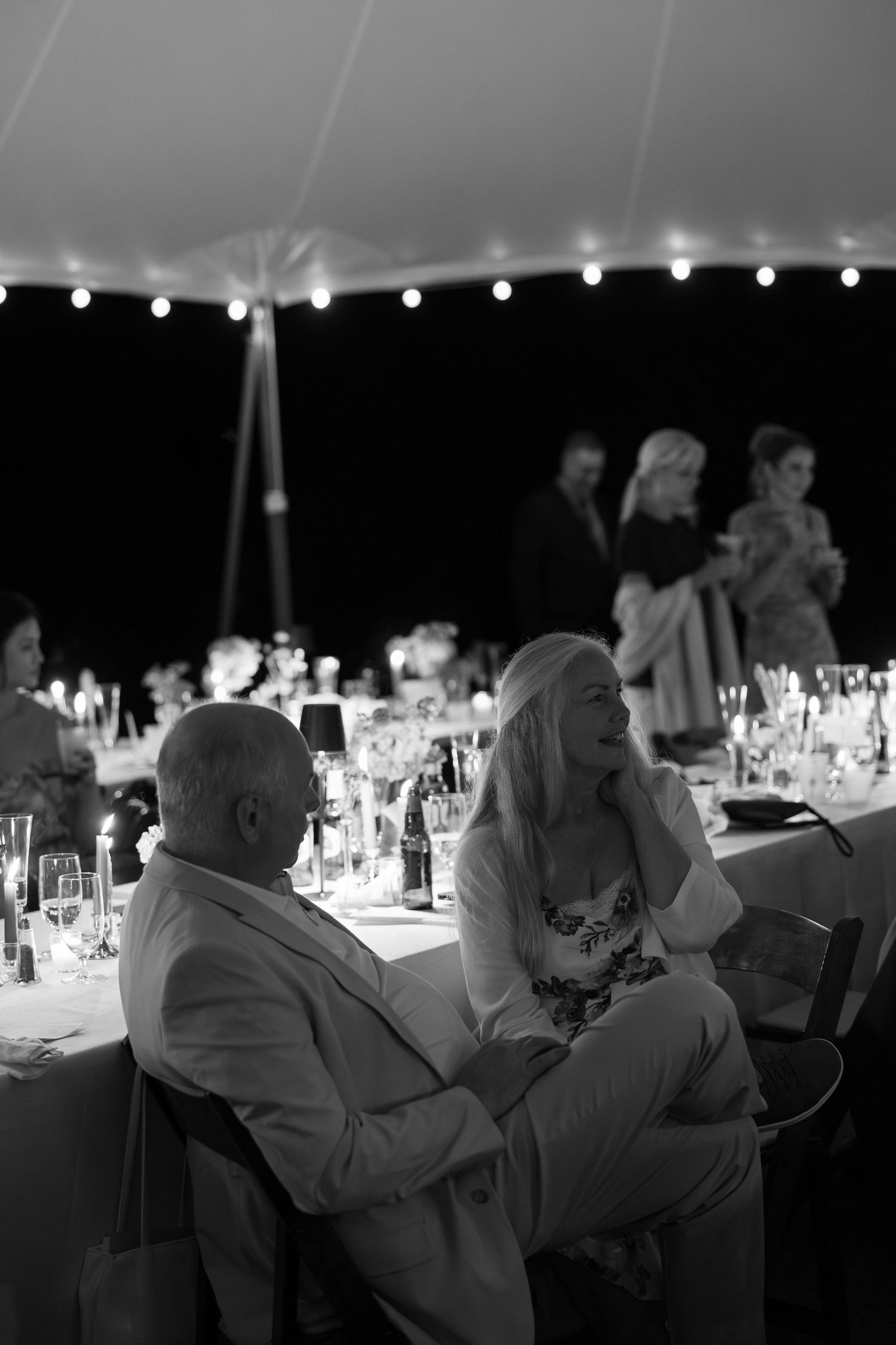 Black and white photo of wedding reception guests relaxing under a tent with string lights at night.