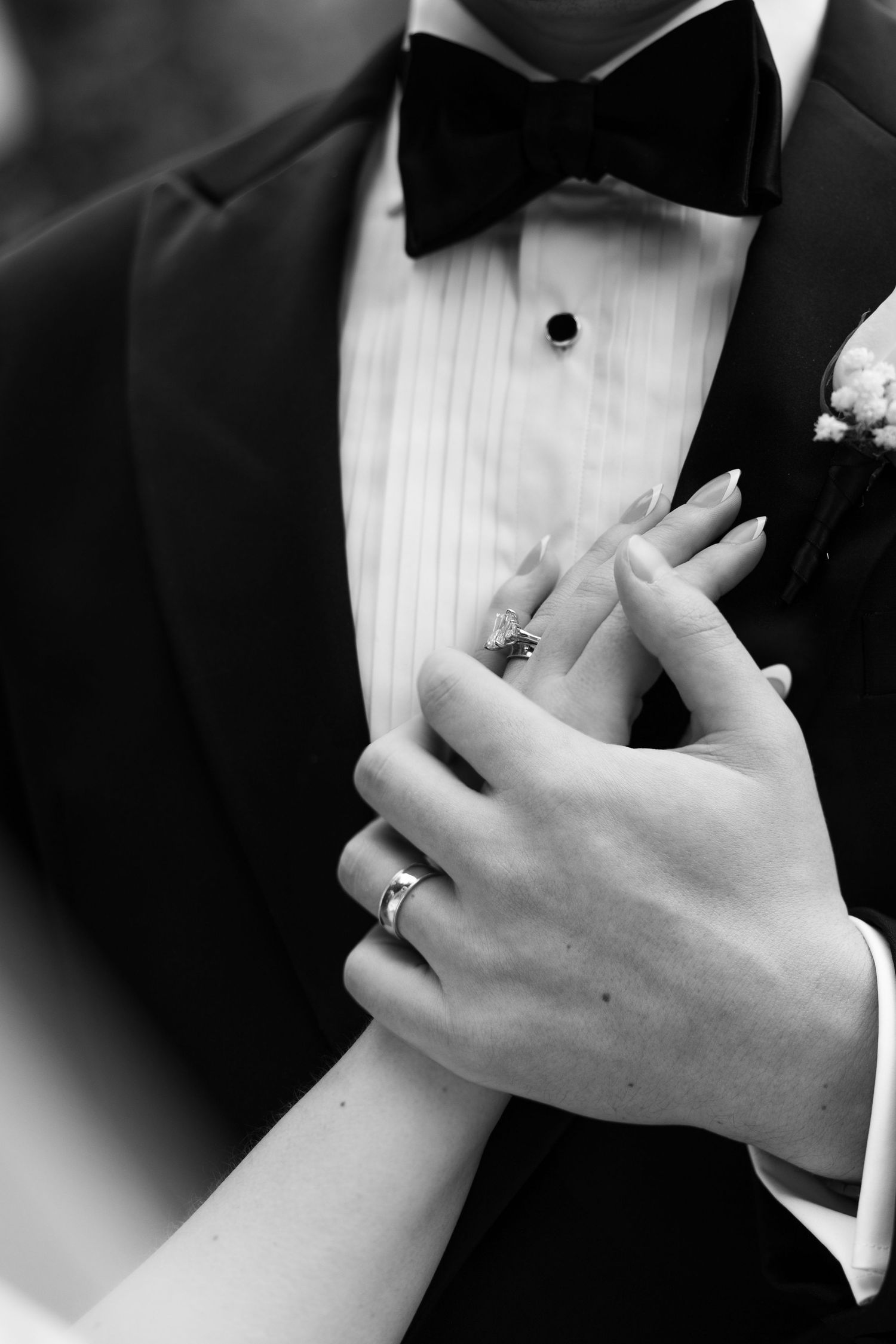 Black and white photo of bride and groom hands with wedding rings, groom in tuxedo with bow tie.