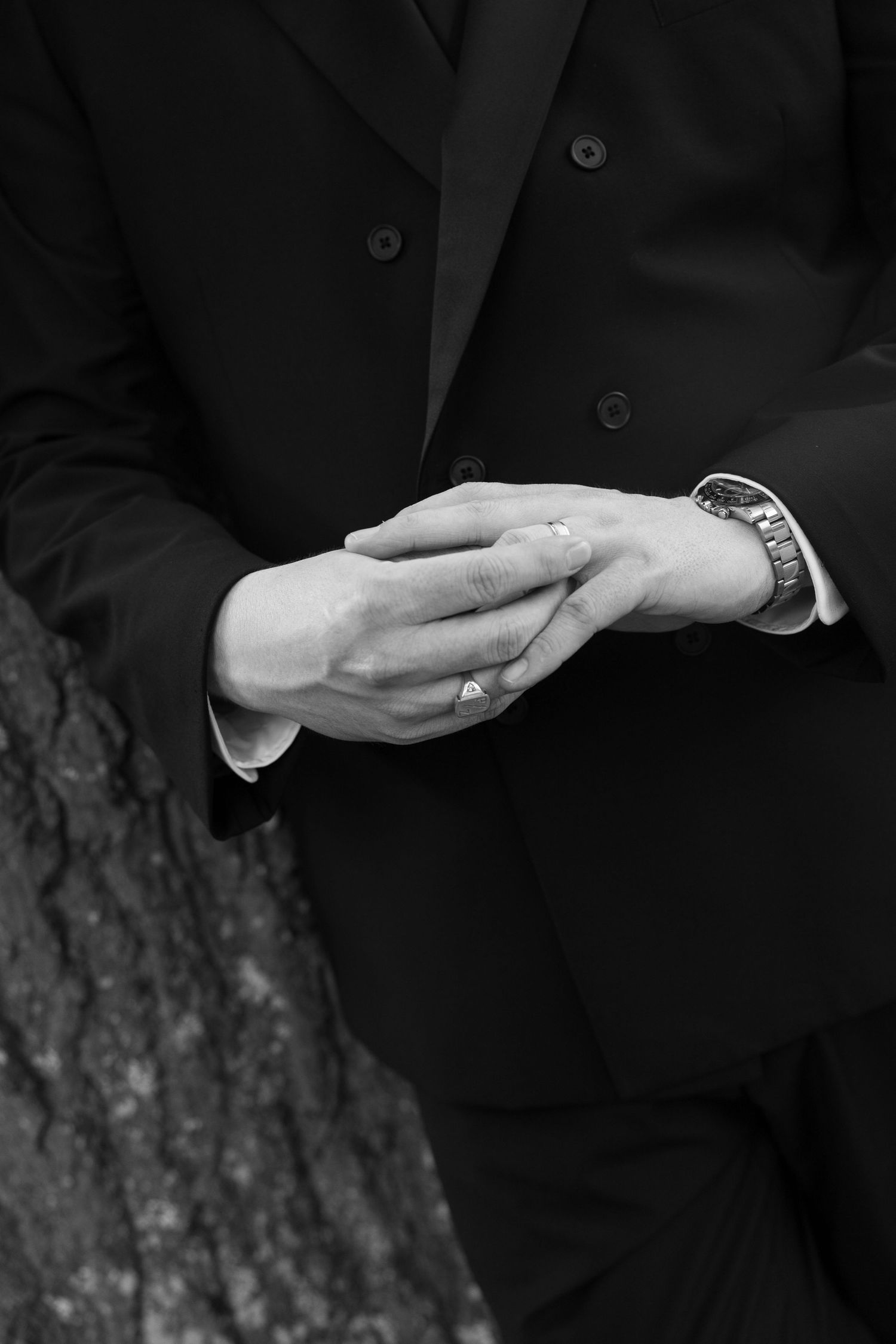 Close-up black and white photo of a groom in a black tuxedo clasping his hands, wearing a watch and wedding ring.