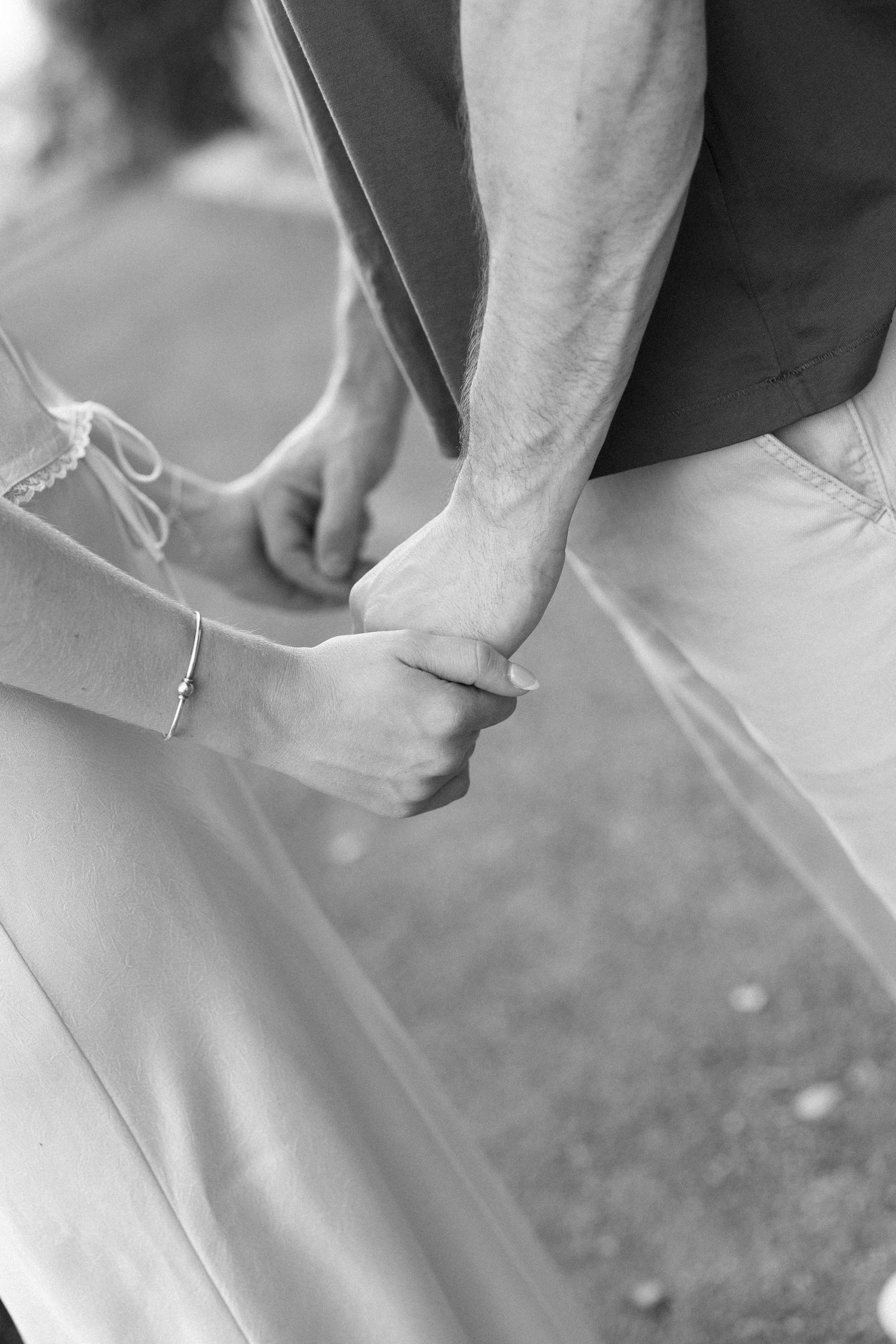Black and white photo of a couple holding hands, bride wearing bracelet, groom in khaki pants, romantic outdoor setting.