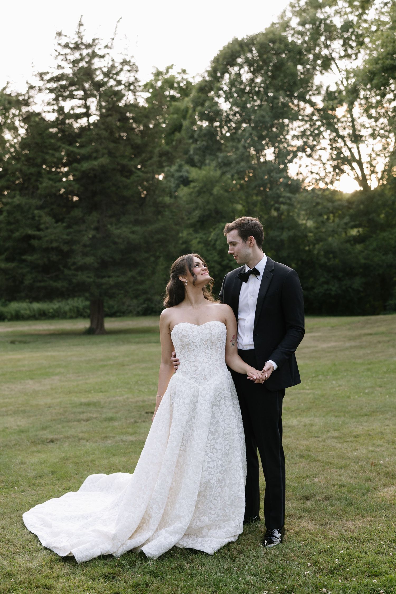 Bride in strapless lace gown and groom in black tuxedo share a romantic moment on green lawn at sunset.
