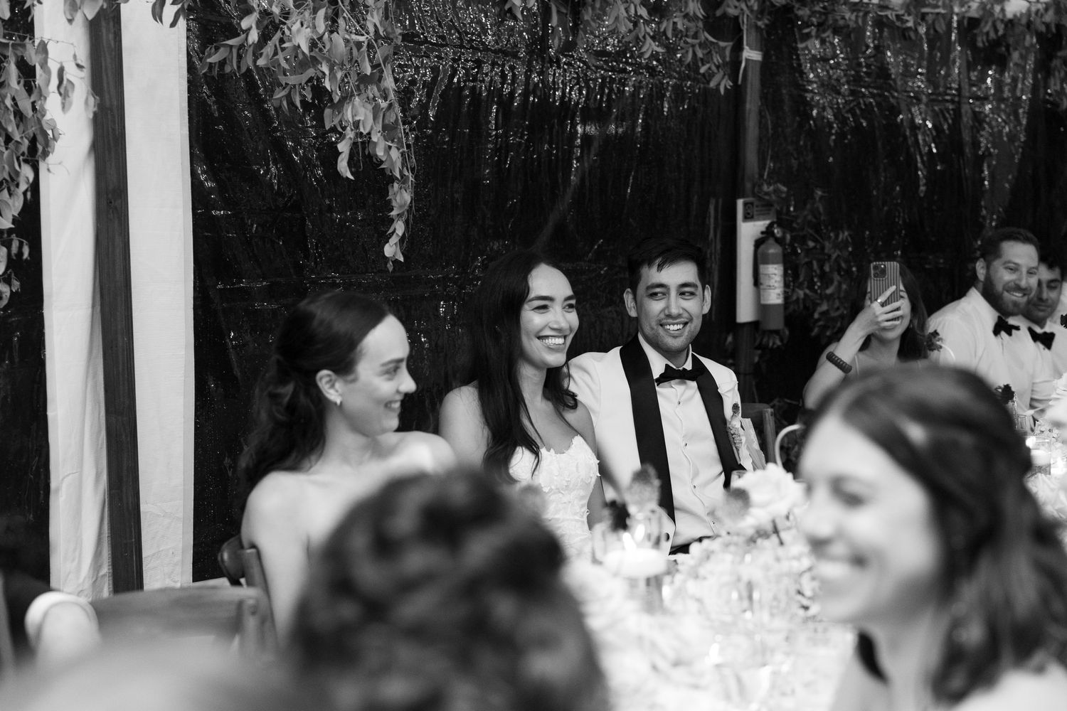 Black and white wedding reception photo of smiling bride and groom seated at head table with guests.