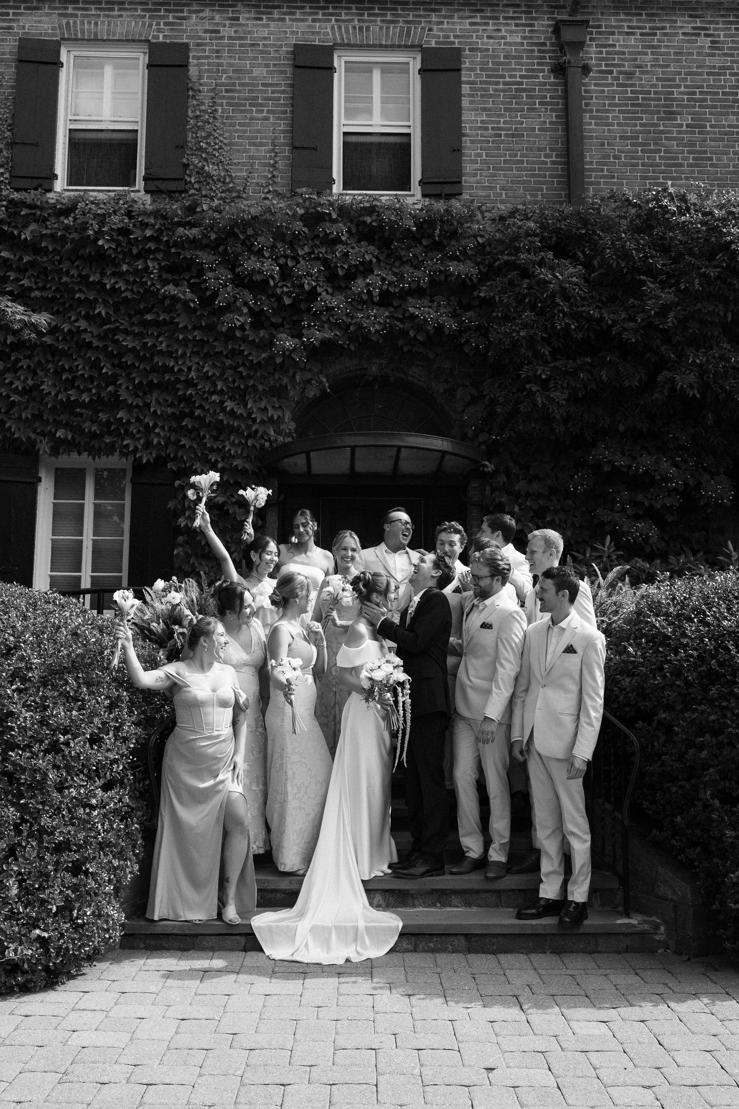 Black and white wedding party photo outside ivy-covered brick building, bride and groom kissing surrounded by bridesmaids and groomsmen.
