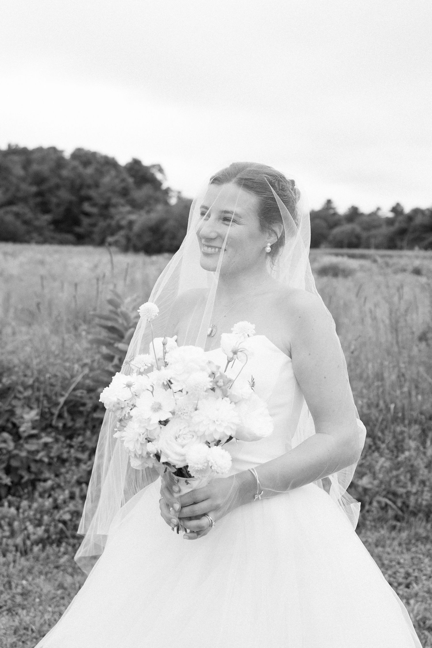 Smiling bride in white gown and veil holds bouquet of white flowers in a scenic outdoor field.