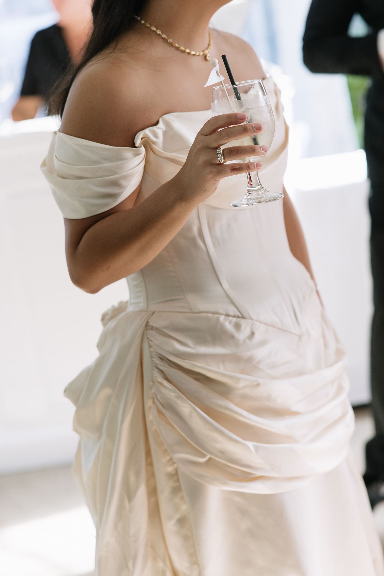 Bride in off-shoulder satin gown holds cocktail wearing pearl necklace.