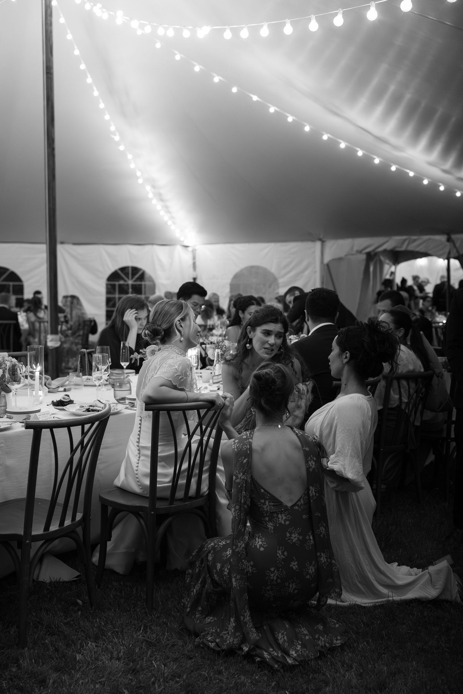 Black and white photo of wedding guests conversing at tables under a string-lit reception tent.
