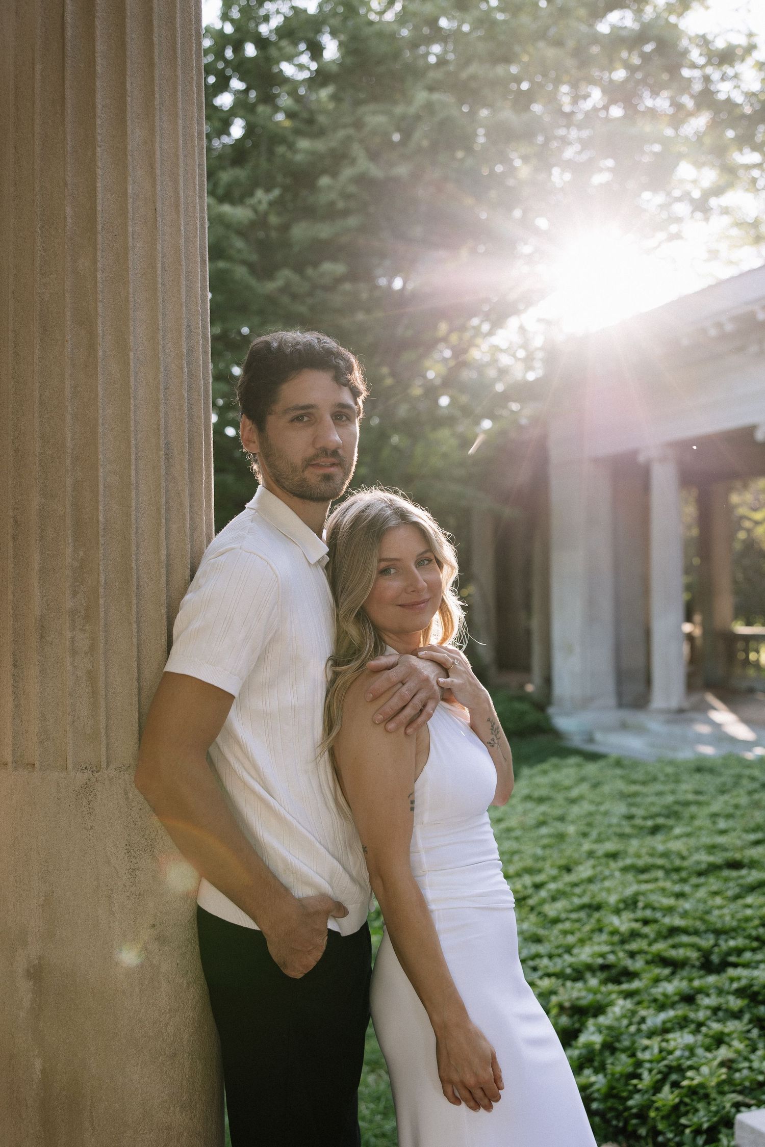 Couple posing by stone column at sunset in lush garden, woman in white dress smiling, man in white shirt embracing her.