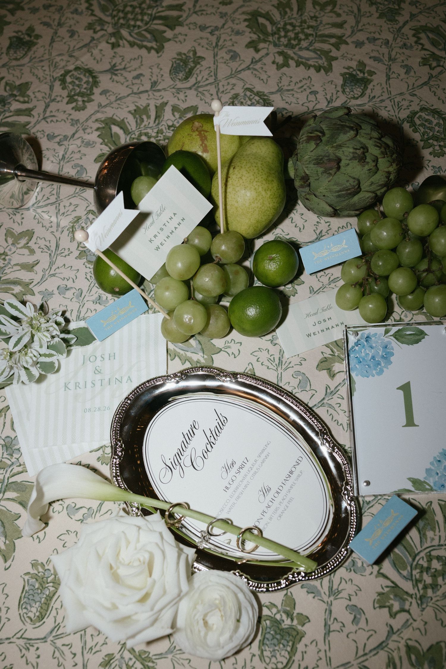 A wedding table displays a silver signature cocktails menu, green apples, grapes, an artichoke, white roses, and escort cards.