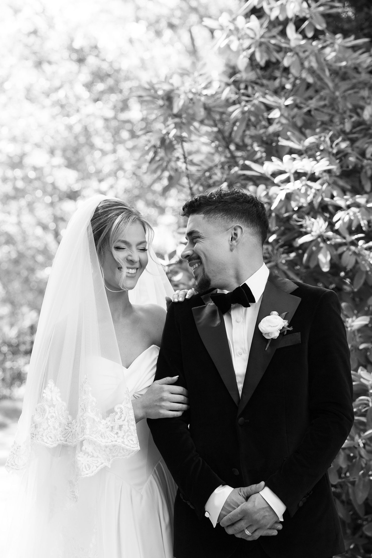 Black and white wedding portrait of a laughing bride in a lace-trimmed veil and a groom in a velvet tuxedo with bow tie among lush greenery.