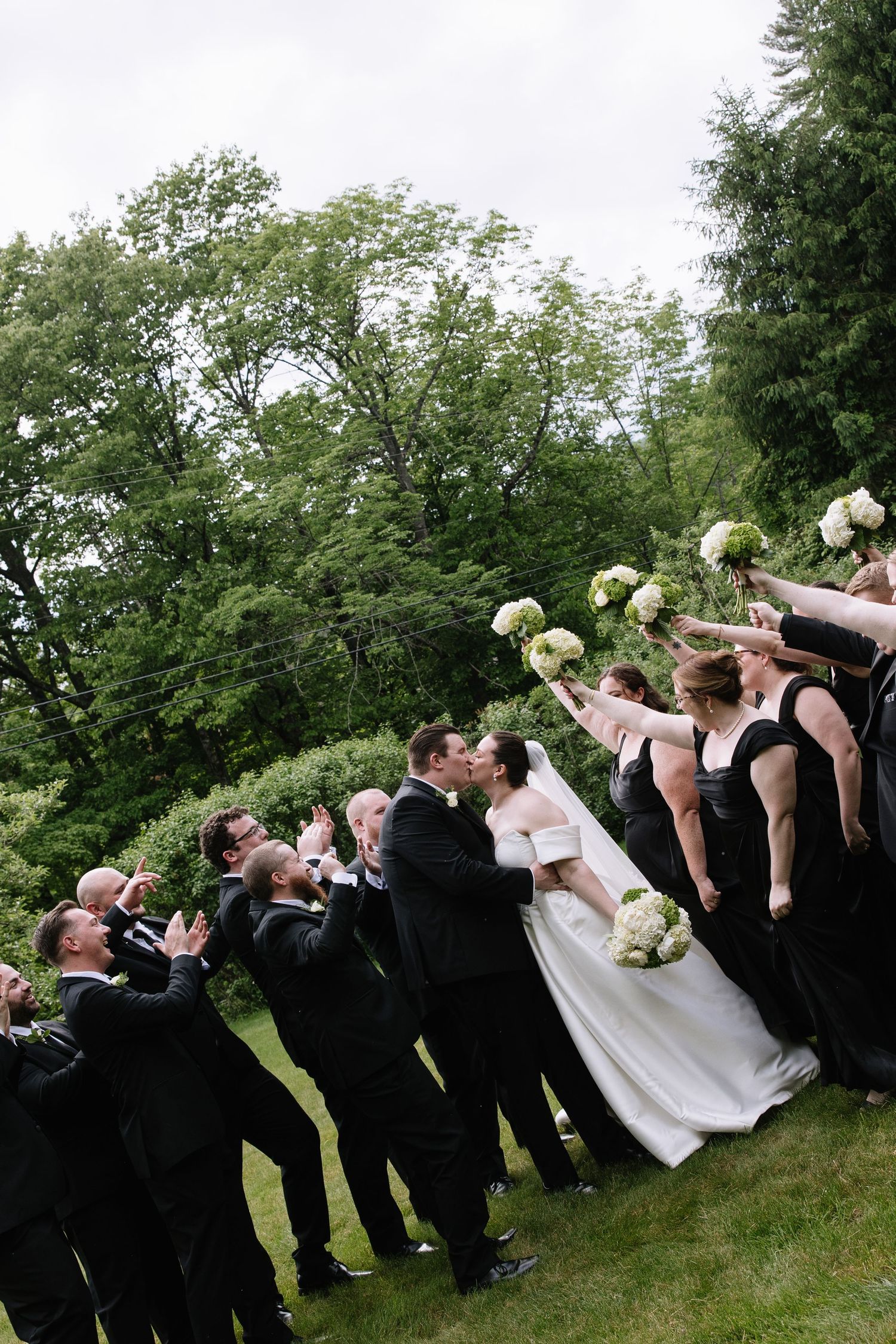 Wedding party celebrates as bride and groom kiss outdoors, bridesmaids in black dresses raise bouquets while groomsmen cheer.