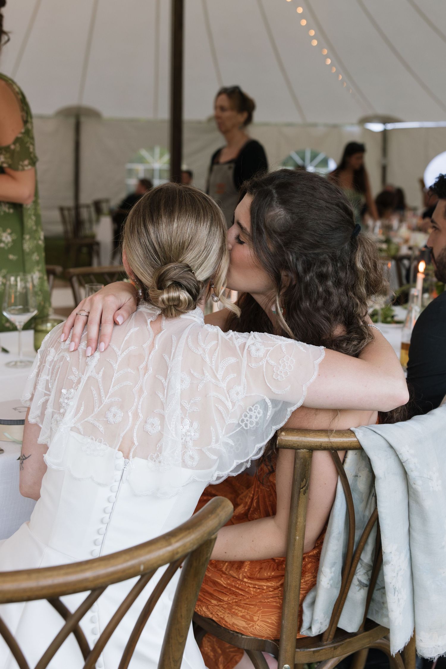 Two women sharing a kiss at wedding reception under white tent, bride in lace dress.