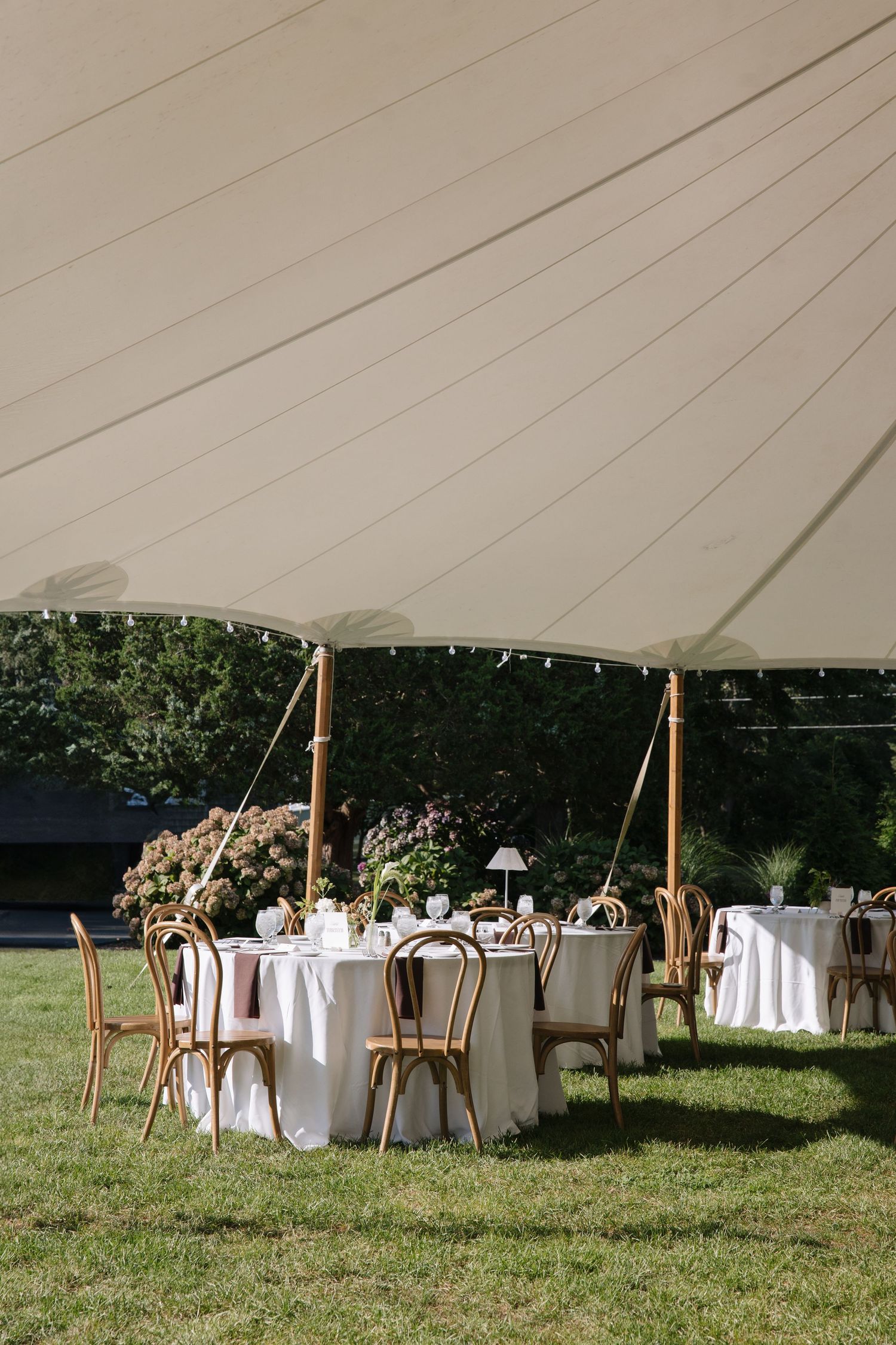 Elegant outdoor wedding reception under a white sailcloth tent with round tables, white linens, and wooden chairs on green grass.