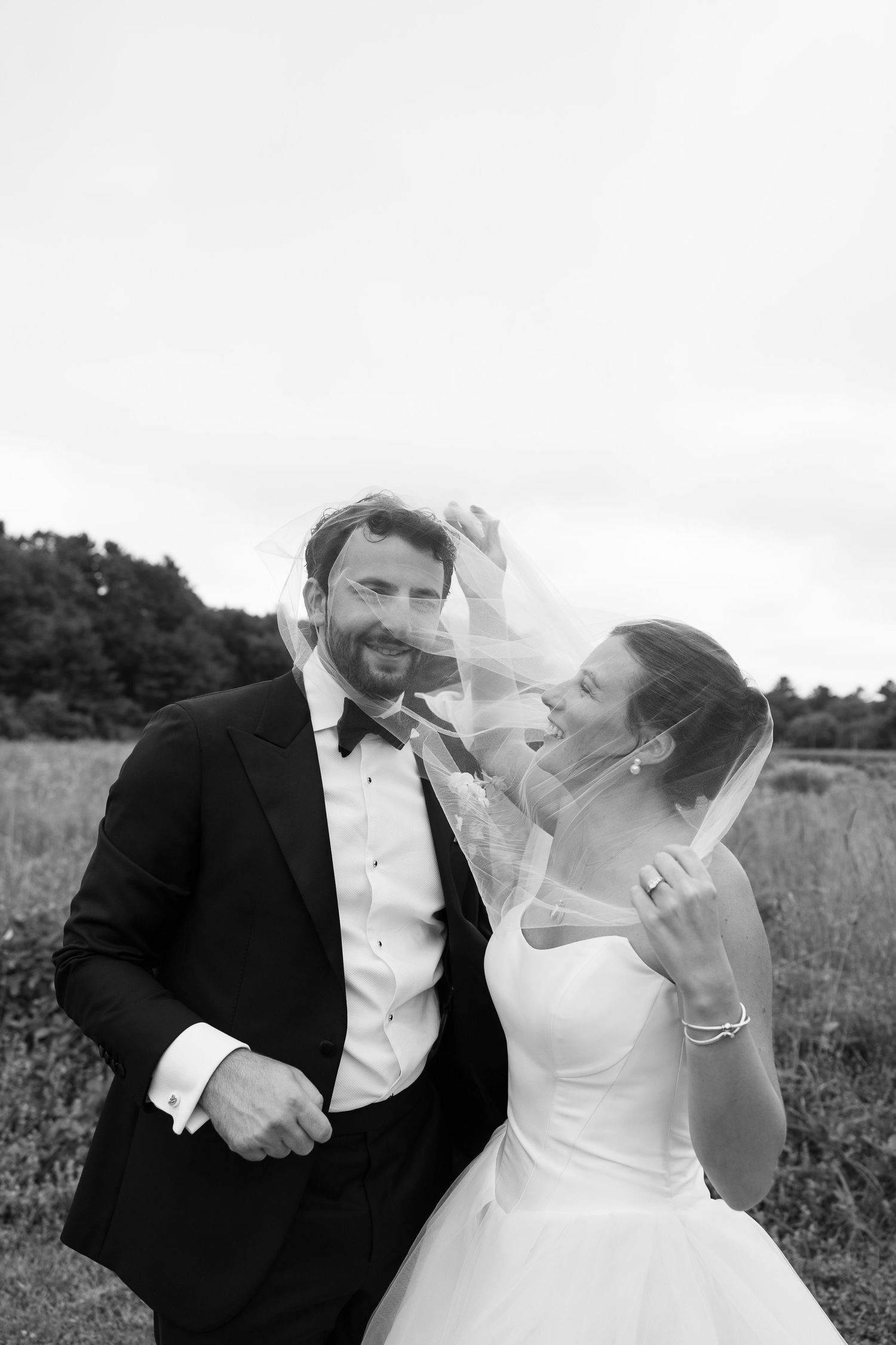 Smiling bride and groom laugh together as veil blows in wind during outdoor wedding portraits.