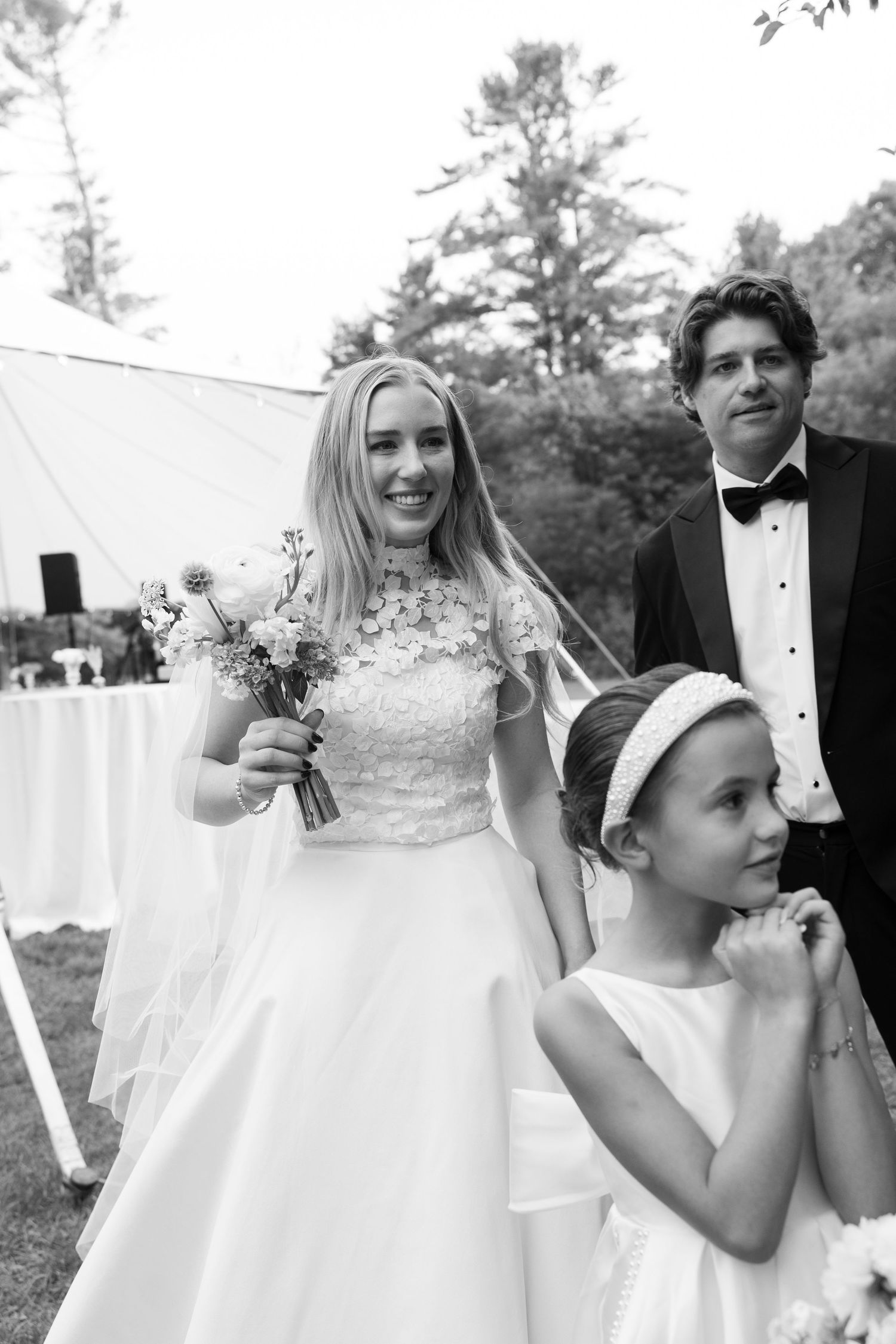 Black and white wedding photo of smiling bride holding bouquet, groom in tuxedo, and flower girl at outdoor ceremony.