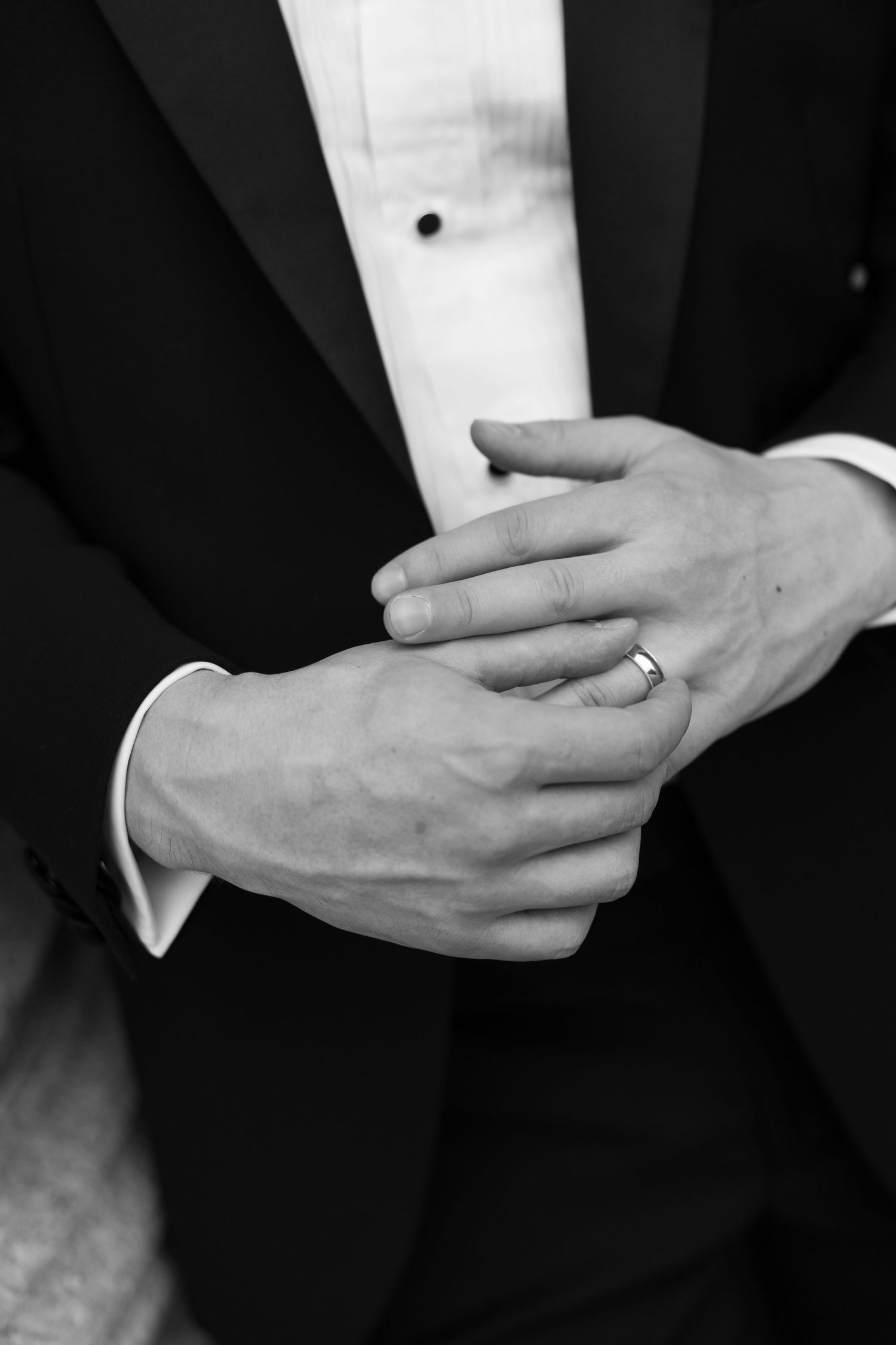 Black and white photo of a groom in tuxedo clasping hands, showing a wedding band ring.