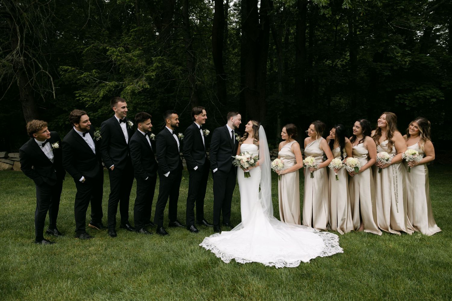 Wedding party posed outdoors with bridesmaids in light dresses and groomsmen in dark suits.