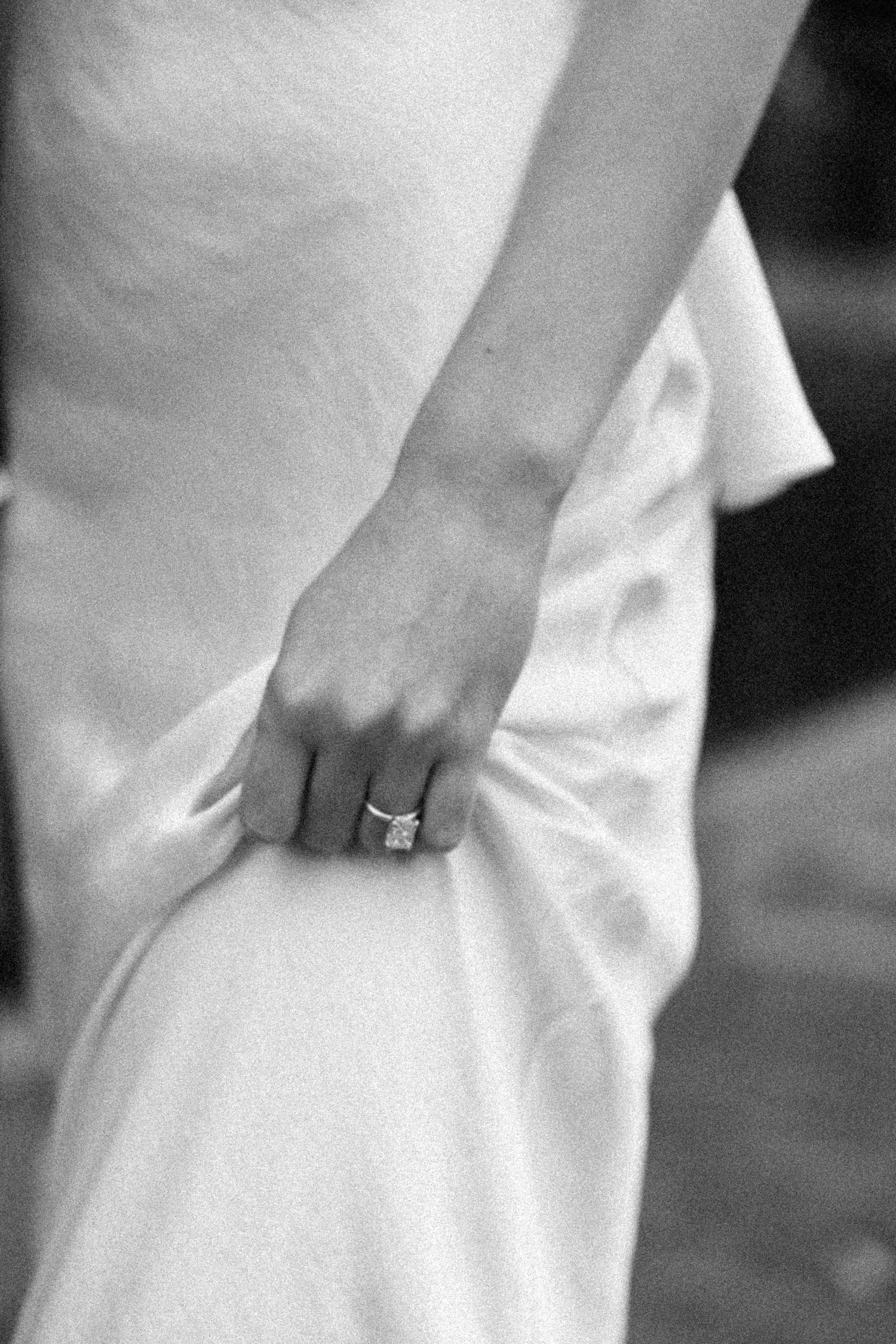 Close-up black and white shot of a hand wearing an engagement ring against a white dress fabric.