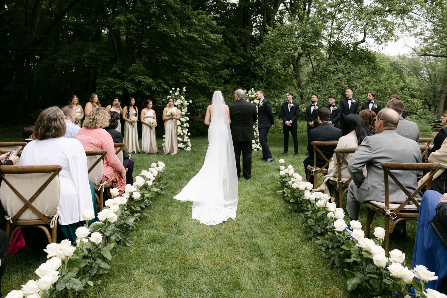 An outdoor wedding ceremony with white flower petals lining the aisle in a wooded setting.