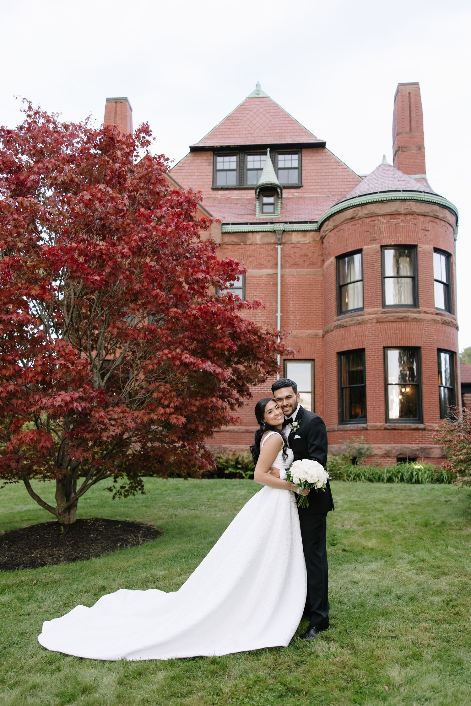 Wedding couple poses in front of a grand brick mansion with vibrant red autumn trees.