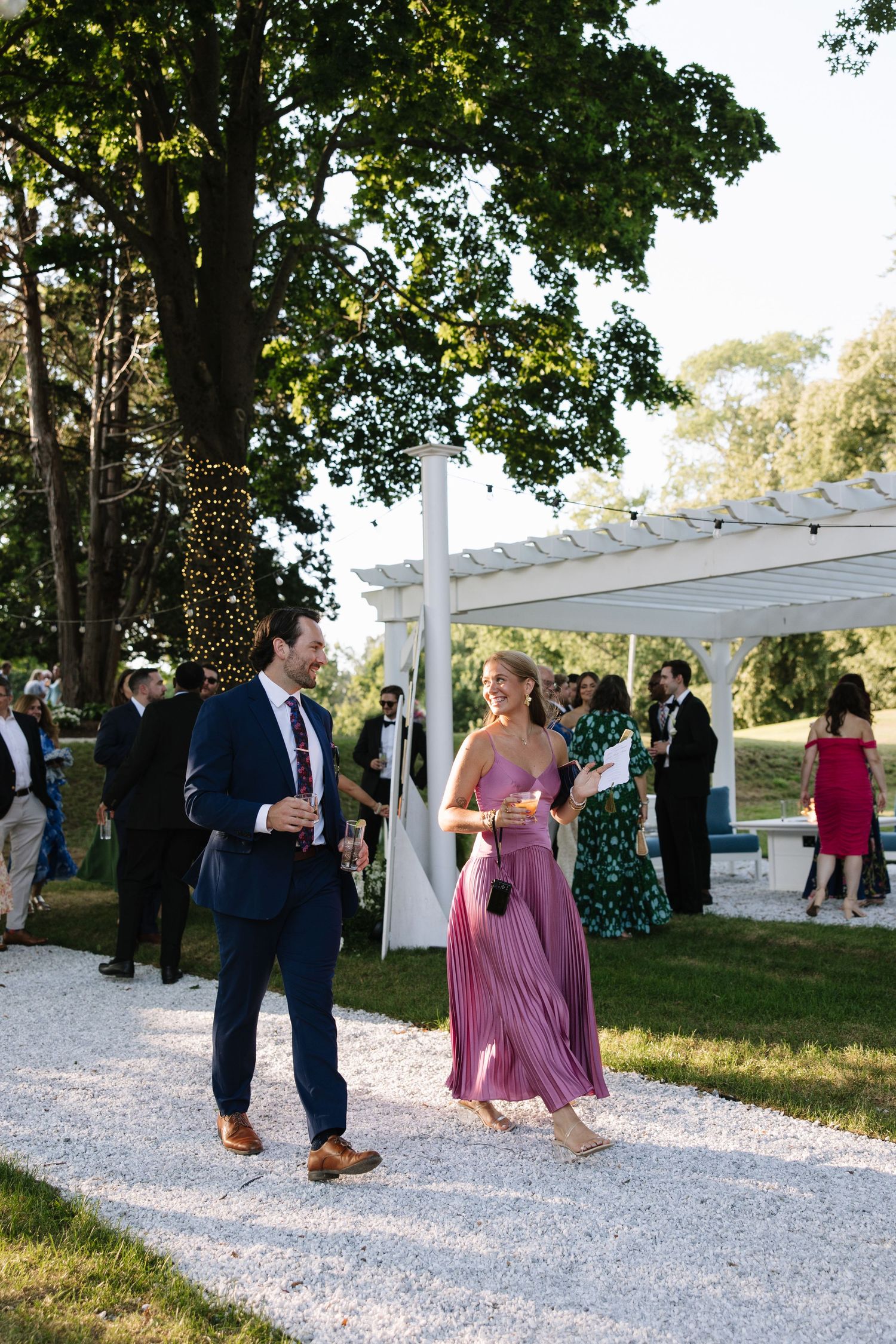 Couple in formal attire walking on gravel path at outdoor wedding reception with white pergola and string lights.