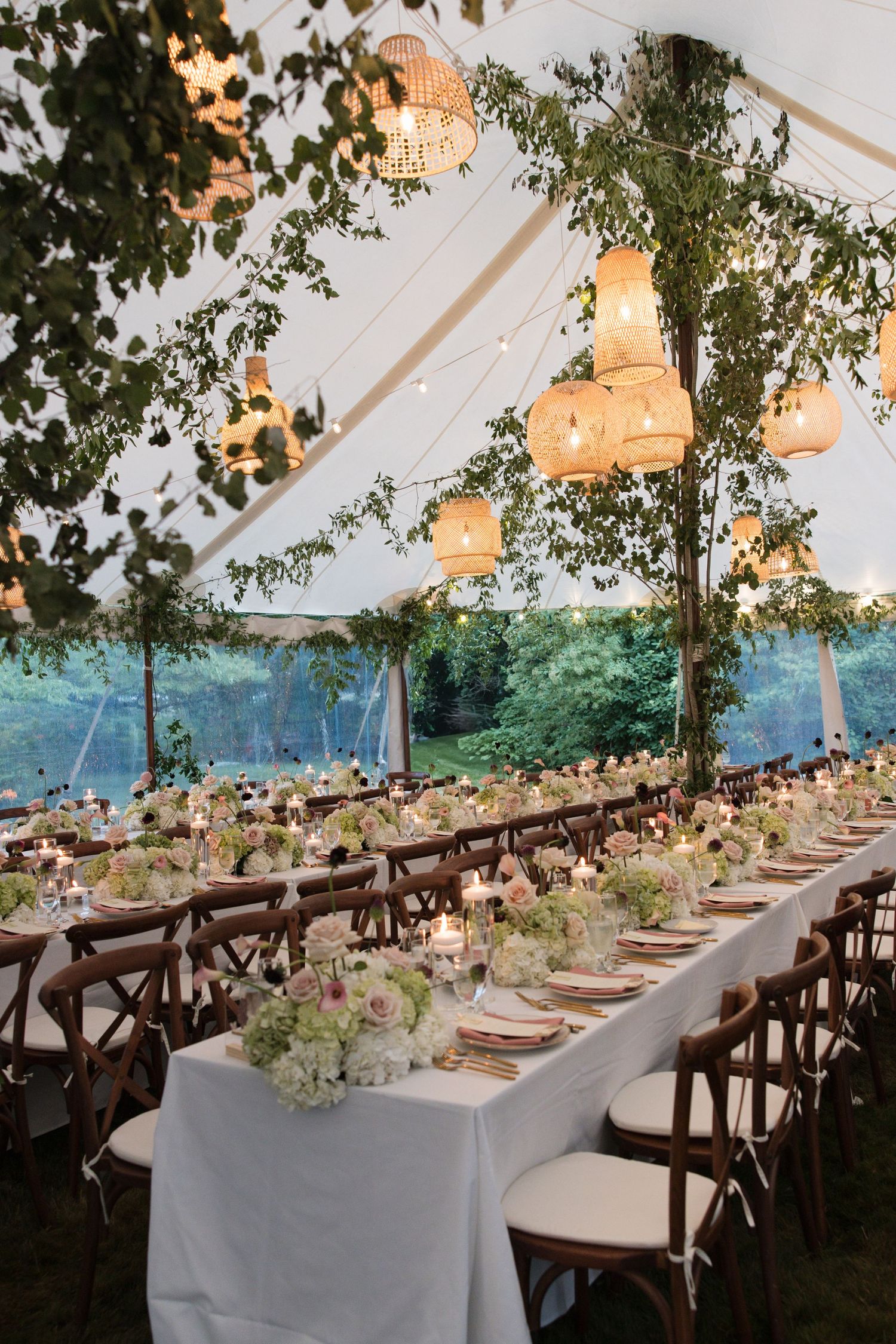 Romantic outdoor wedding tent reception with long tables, floral centerpieces, candles, and wicker pendant lanterns at dusk.