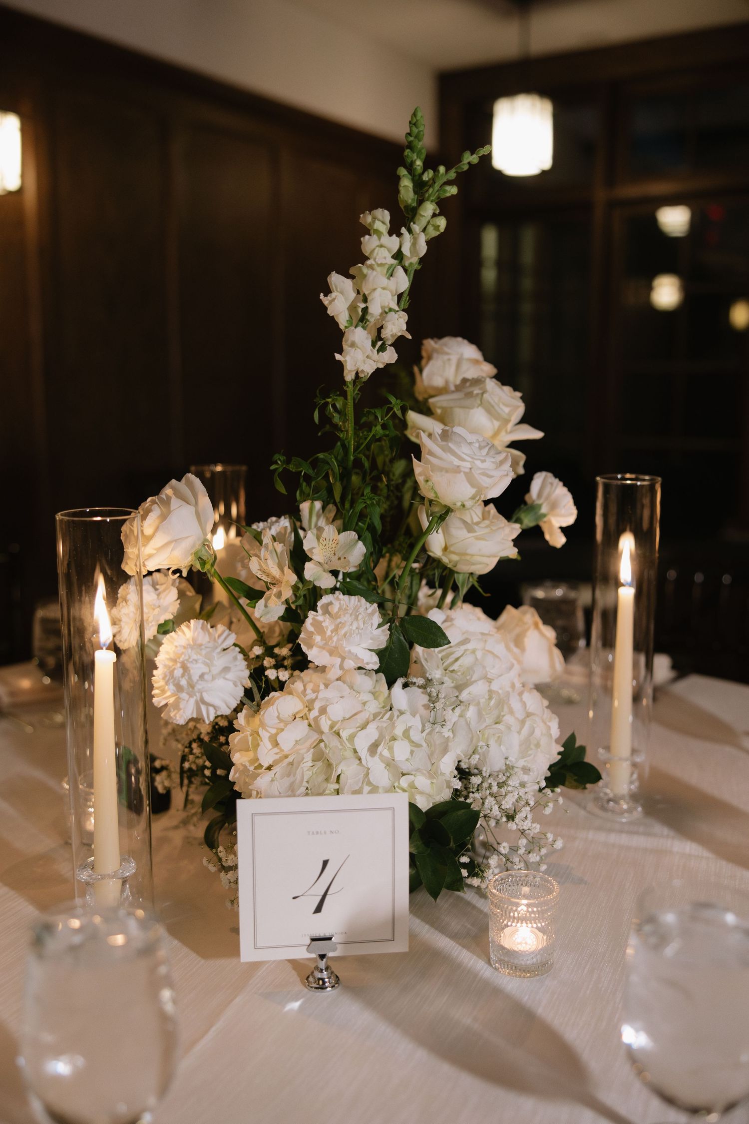 An elegant wedding reception table decorated with white flowers, candles and greenery on a white tablecloth.