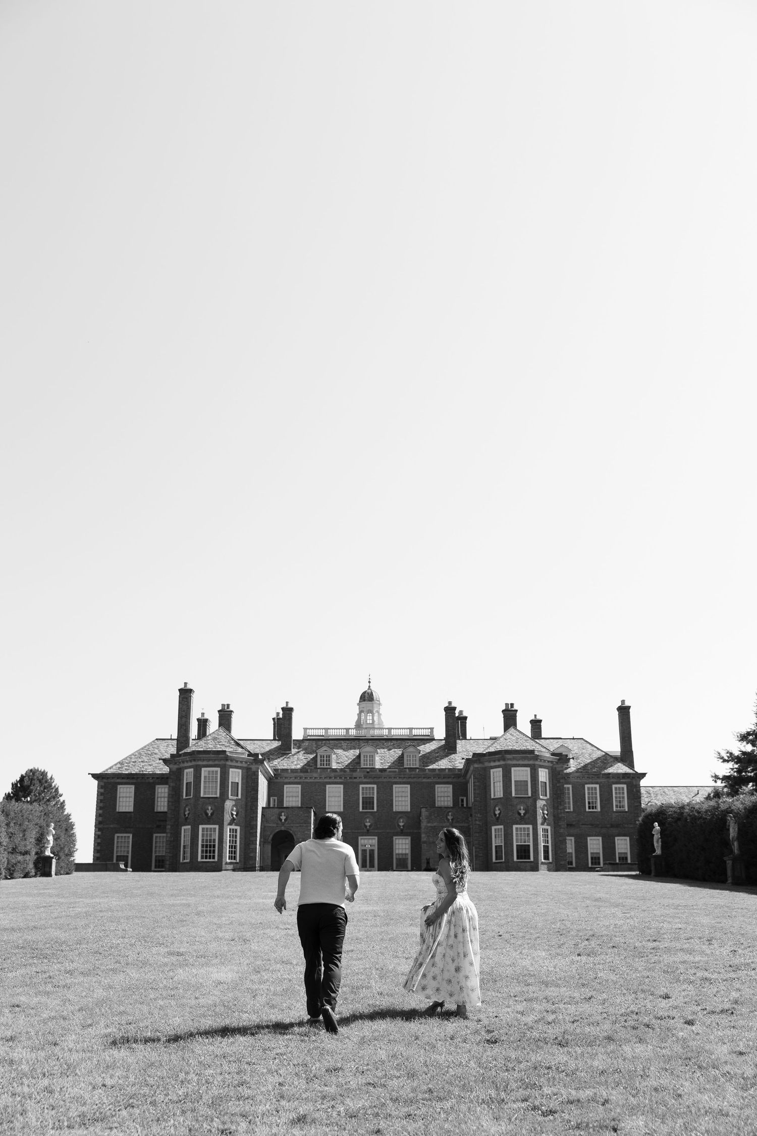 Black and white photo of a couple holding hands walking toward a grand historic manor house across a vast lawn.